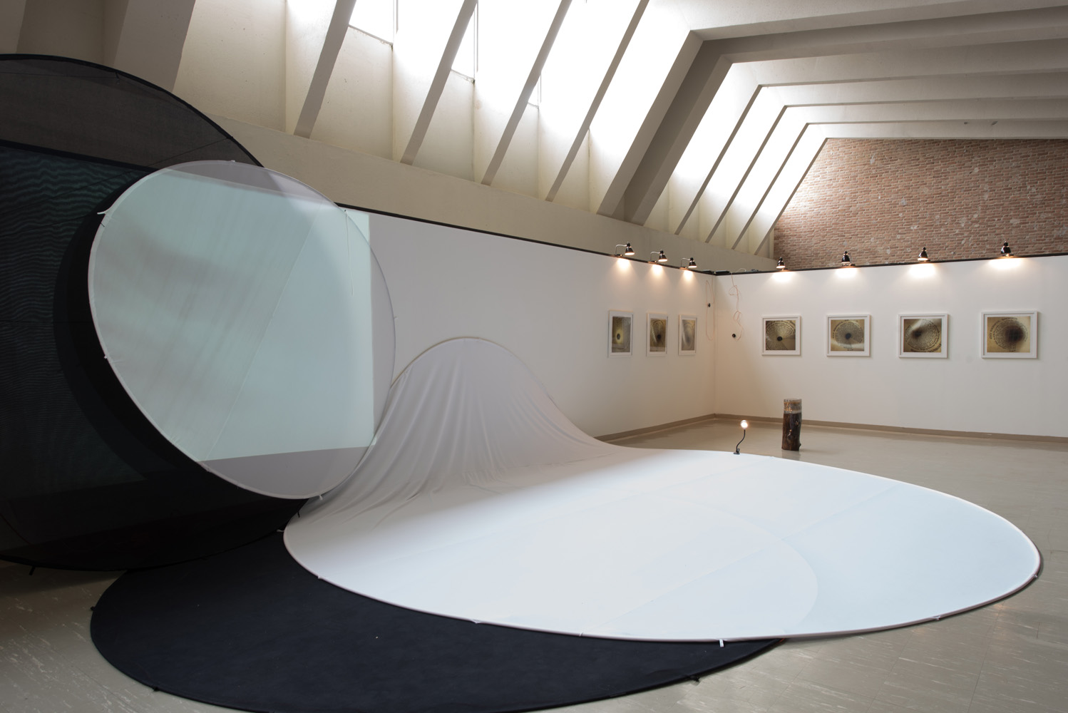 Indoor art gallery featuring a modern installation with large black, white, and semi-transparent fabric in flowing shapes. Framed artworks are displayed on the white wall, illuminated by spotlights. The gallery has a high, angled ceiling with skyligh