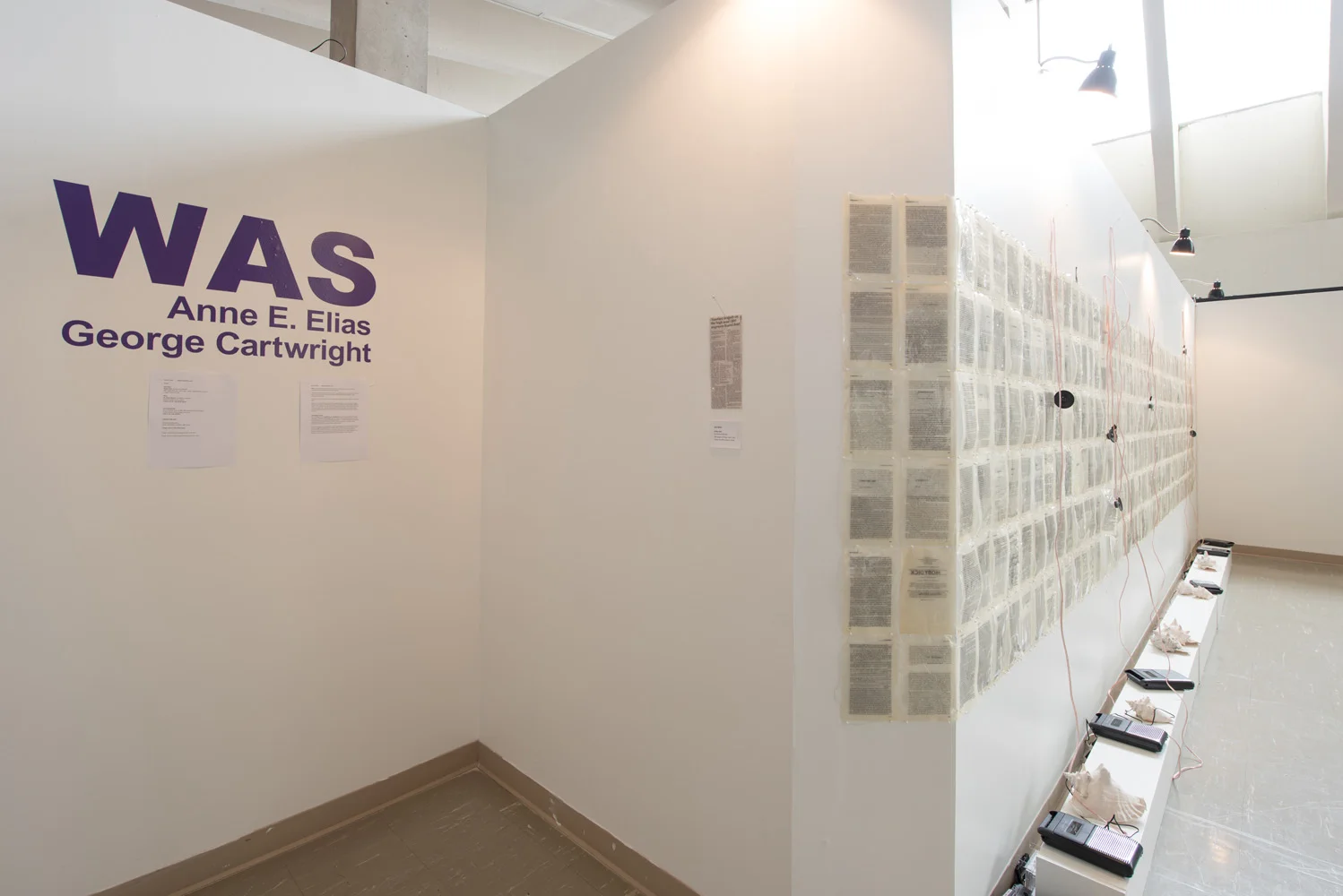 Exhibition wall titled 'WAS' by Anne E. Elias and George Cartwright, displaying numerous pages printed on paper, connected with red wires, and accompanied by clipboards and small objects on a ledge.