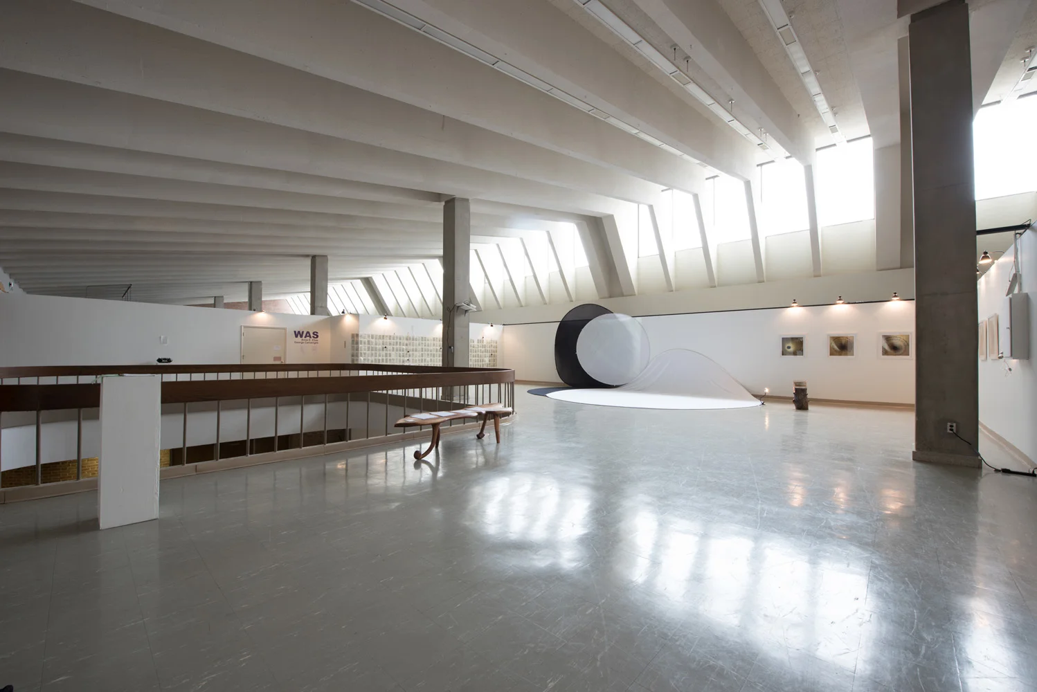 Modern art gallery interior with large black and white spherical sculptures, small framed artworks on the wall, and a bench near a railing, with bright natural light from windows above.