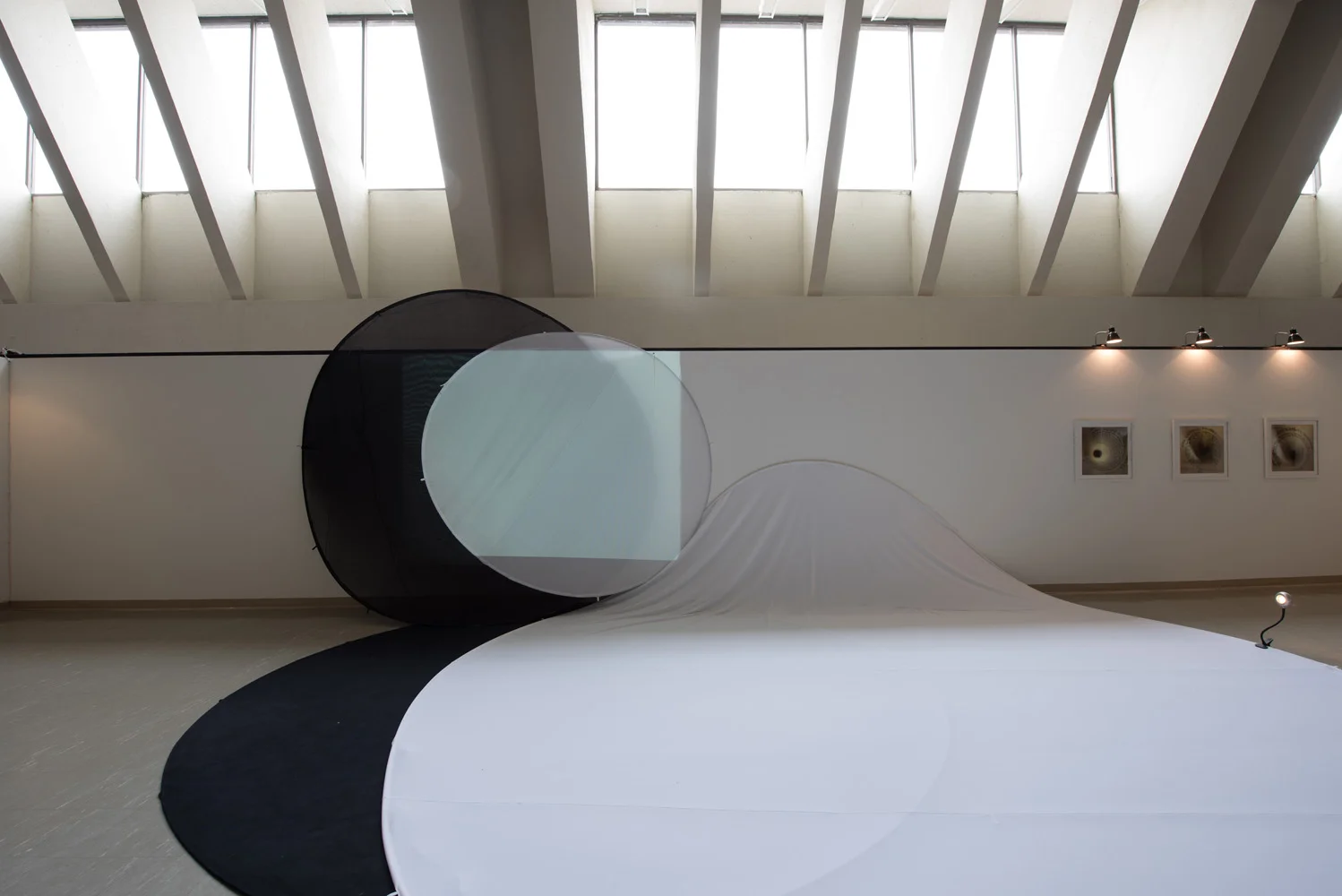 Contemporary art installation featuring a large white fabric draped over a surface with black circular structures and a projection screen, in an art gallery with skylights and framed photographs on the wall.