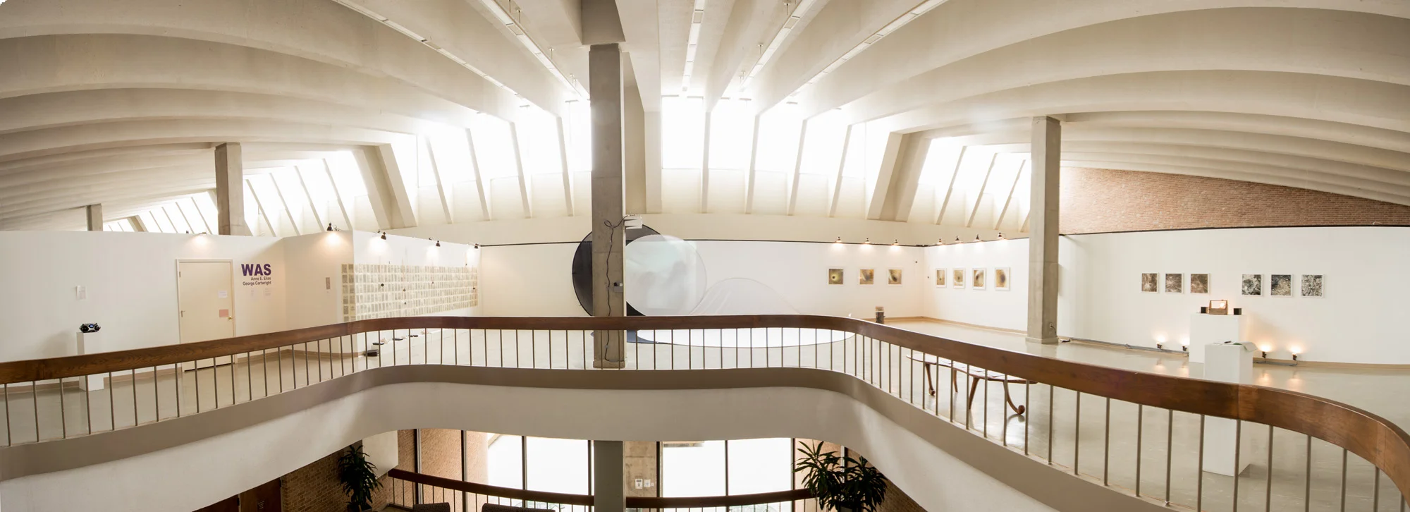 Interior view of an art gallery with a curved balcony, white walls displaying framed artwork, and a high, vaulted ceiling with large windows letting in natural light.