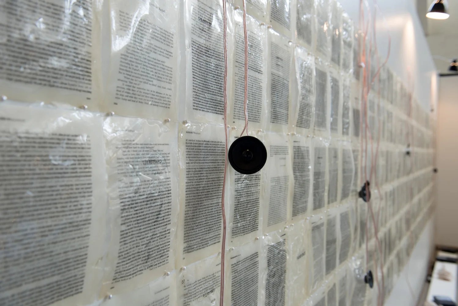 A wall covered with numerous sheets of printed text and electronic components, including black speakers and red wires, displayed in an exhibit or research setting.