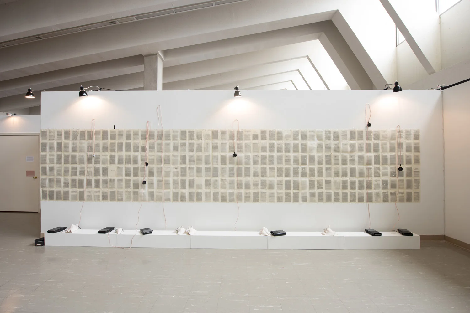 An art installation of numerous pages with text arranged on a large white wall, with black portable speakers and white fabric pieces at the bottom, and electrical cords hanging from the top, illuminated by spotlights in a gallery setting.