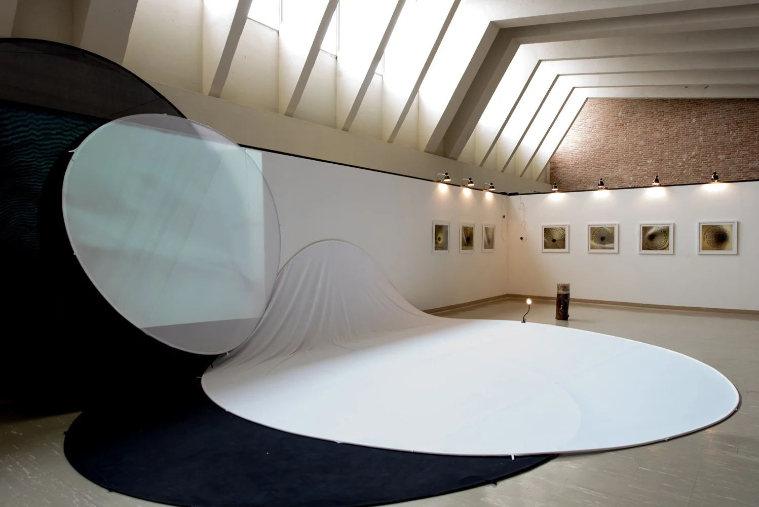 Art installation in gallery with a white fabric sheet draped on a curved structure, and framed artwork on the wall behind. The ceiling has angled windows allowing natural light to illuminate the space.