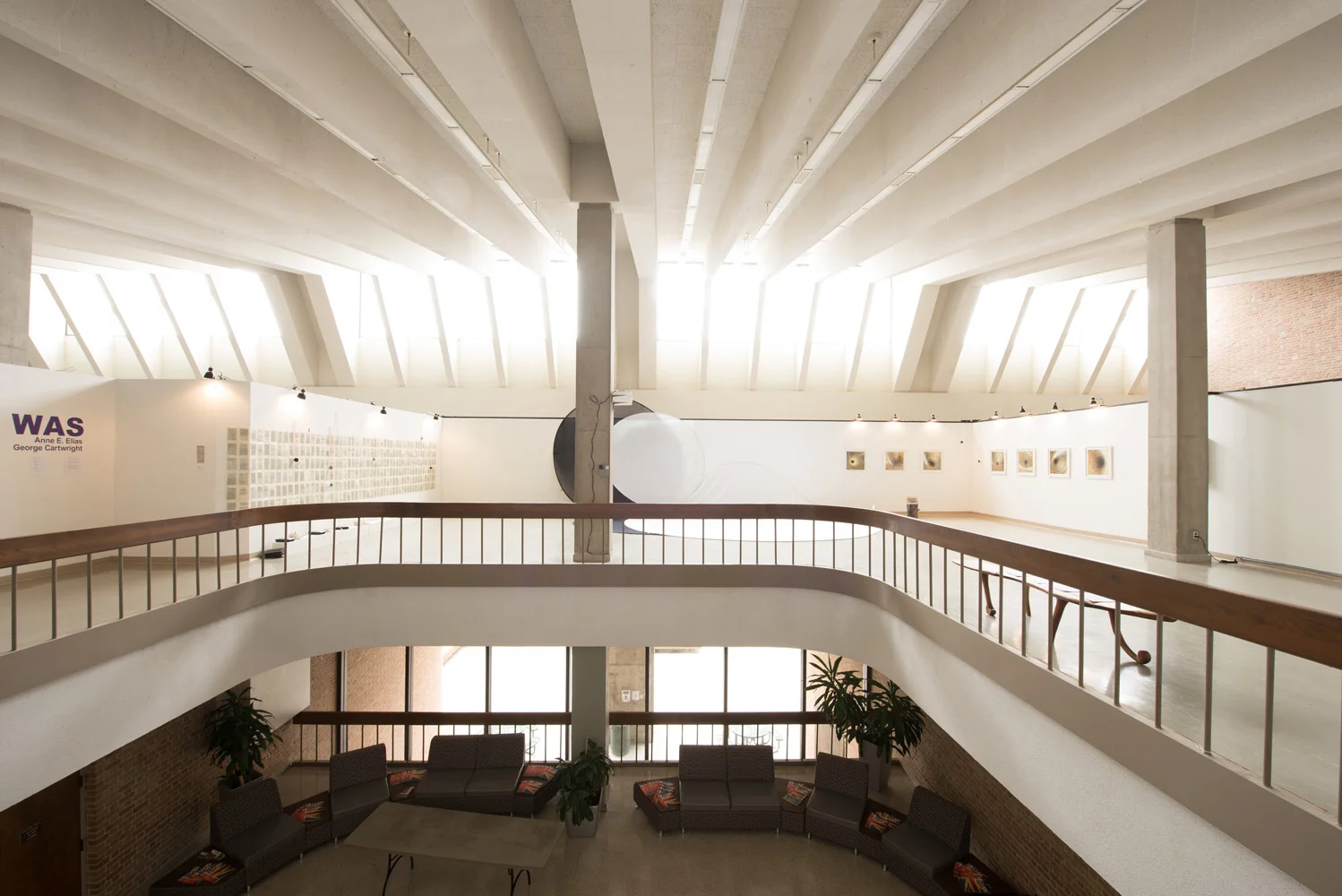 Interior view of a modern art gallery with upper and lower floors, concrete columns, large windows allowing natural light, and artworks on display.