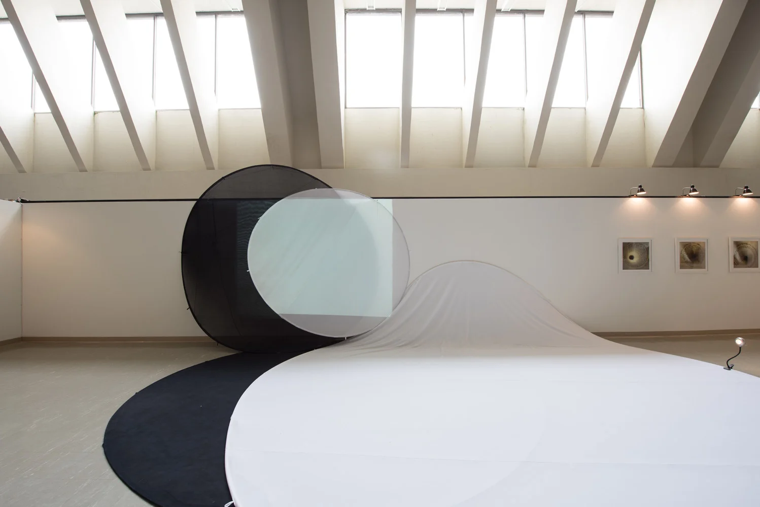 Modern art installation with a black circular frame, a semi-transparent white circle, and a flowing fabric or paper surface in an art gallery with skylight windows.