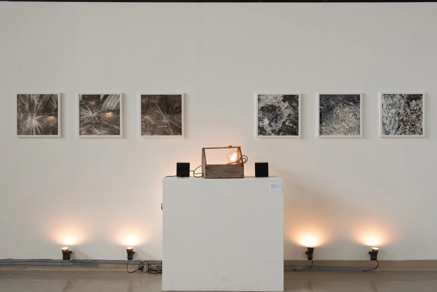 An art gallery display featuring a white wall with six framed black and white photographs, three on each side. In front, there is a white pedestal with a wooden box containing a glowing light bulb and two small black speakers, illuminated by floor sp