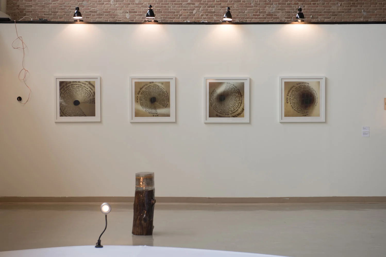 Art gallery exhibit with four framed art pieces on a white wall, a small light on a stand, and a wooden sculpture in the foreground.
