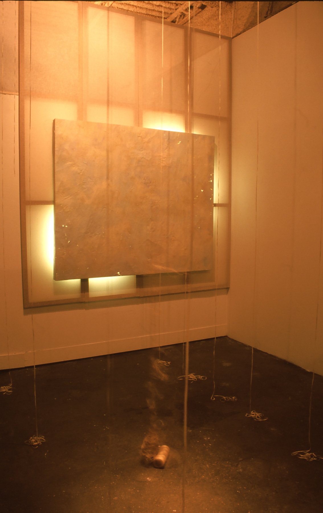 Art installation featuring a large, textured rectangular panel lit from behind, with thin vertical strings hanging from the ceiling, and a small object on the floor.