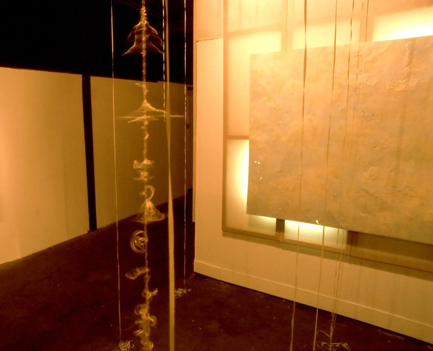 An art installation featuring vertical yellowish strings hanging in front of a textured tile wall with backlighting, and a reflective black surface on the left side reflecting part of the installation.