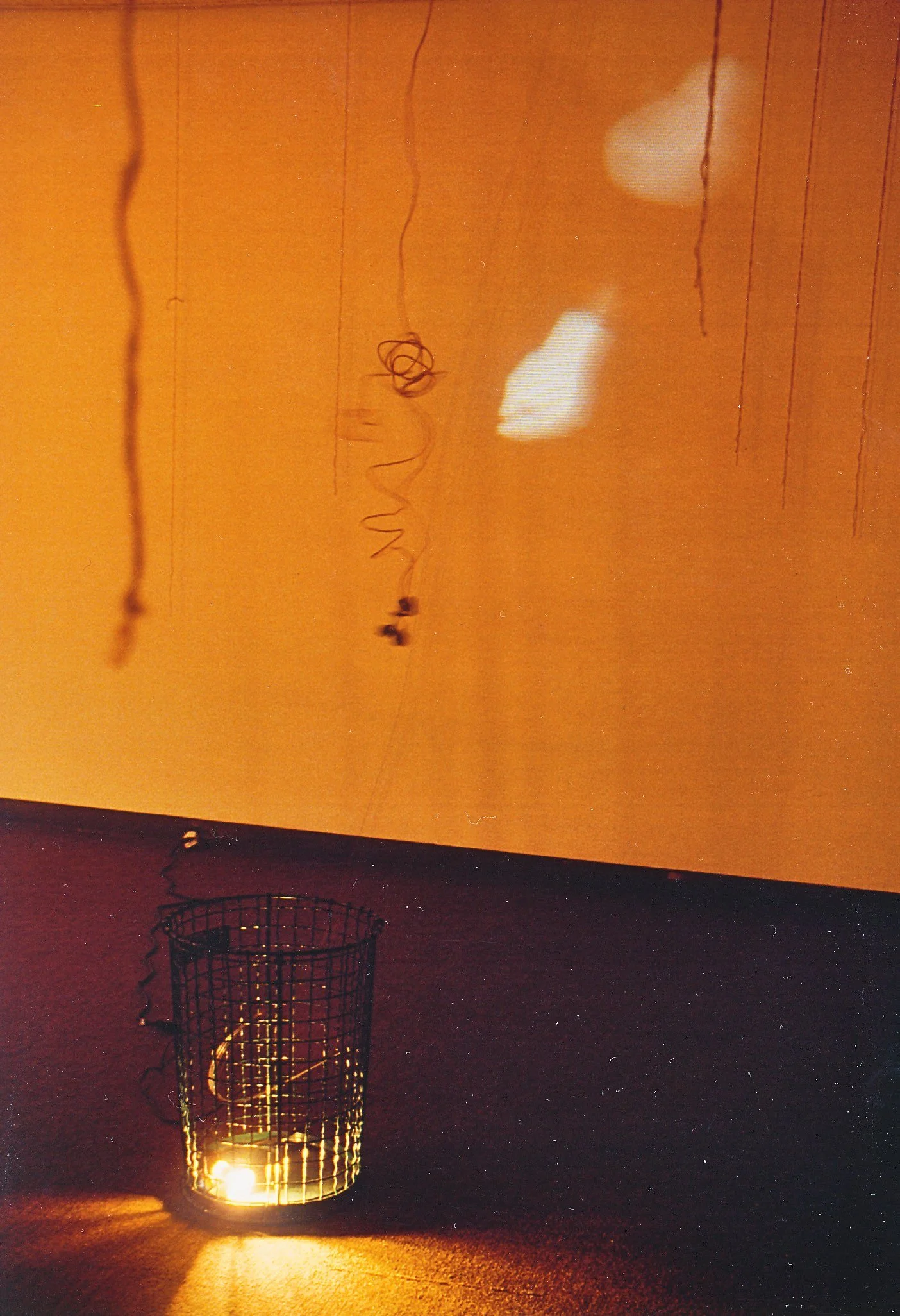 A lit candle inside a wire mesh holder on a dark surface, with a plain orange wall in the background. Shadows and warm light are cast on the wall.