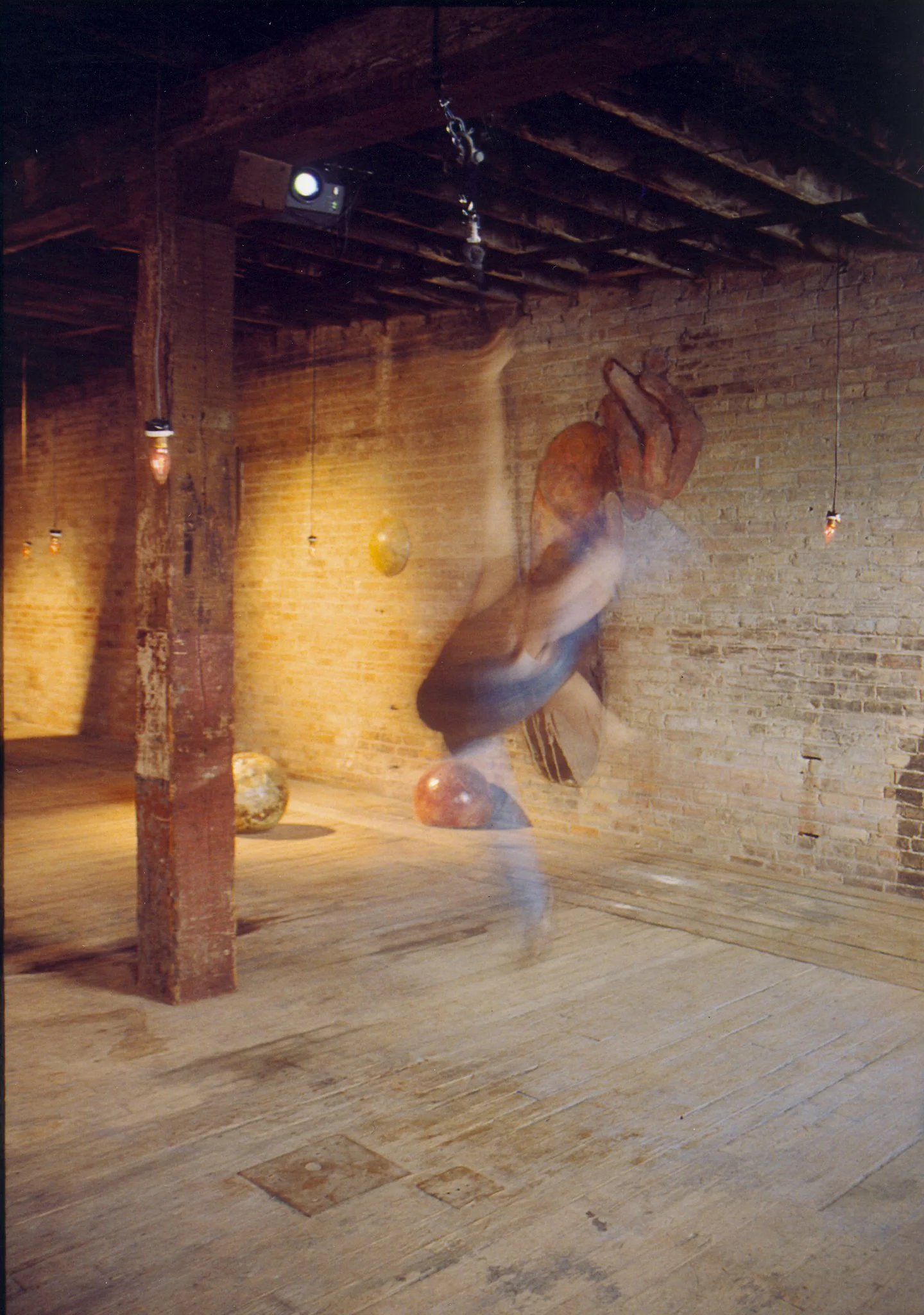 Gallery with wooden floor and brick walls, featuring large hanging sculptural art and spherical objects, illuminated by hanging light bulbs.