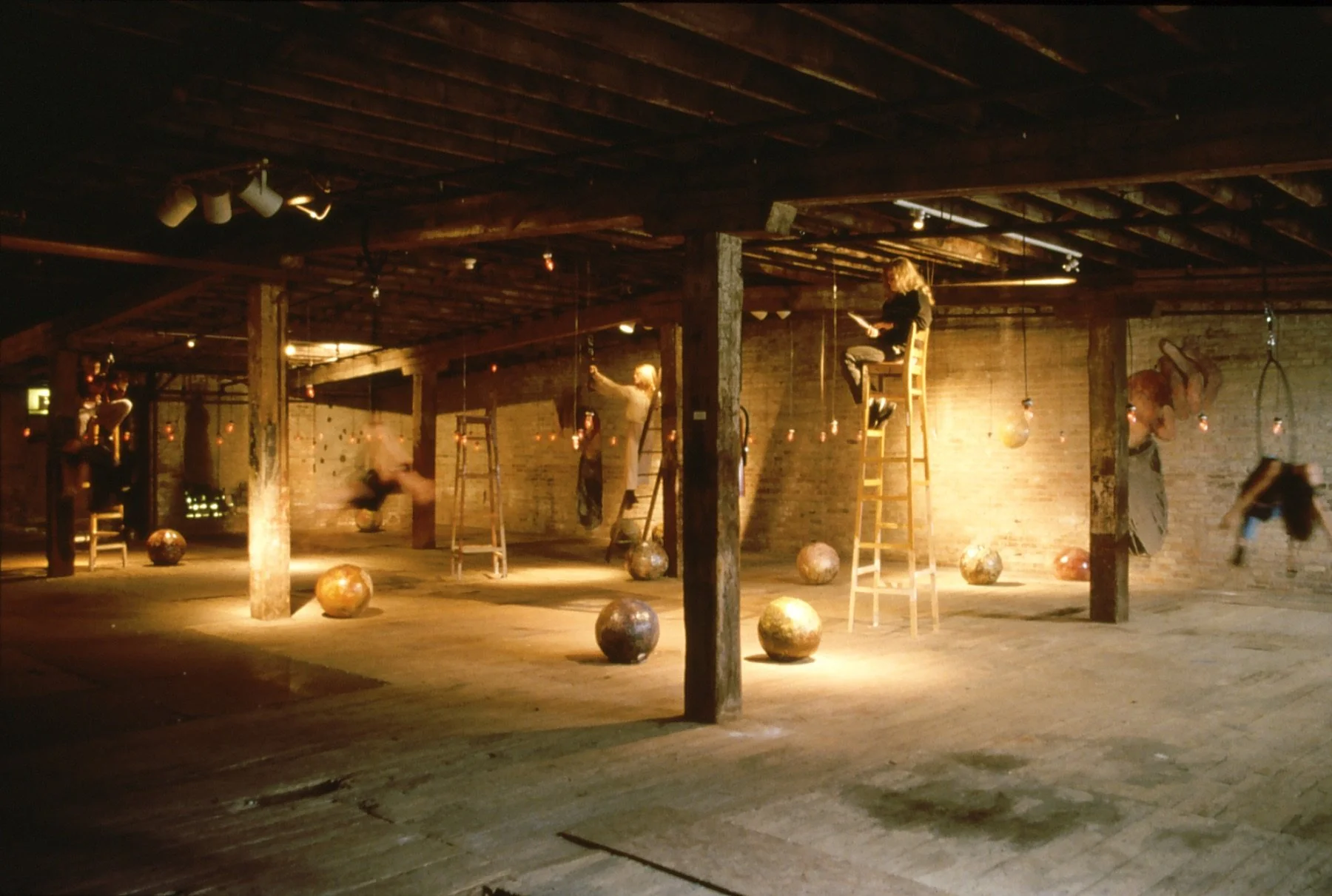 An industrial-style room with exposed brick walls and wooden beams in the ceiling. Several people sitting on ladders reading or working, surrounded by hanging objects and large metallic orbs on the floor. Warm, ambient lighting creates a cozy atmosph