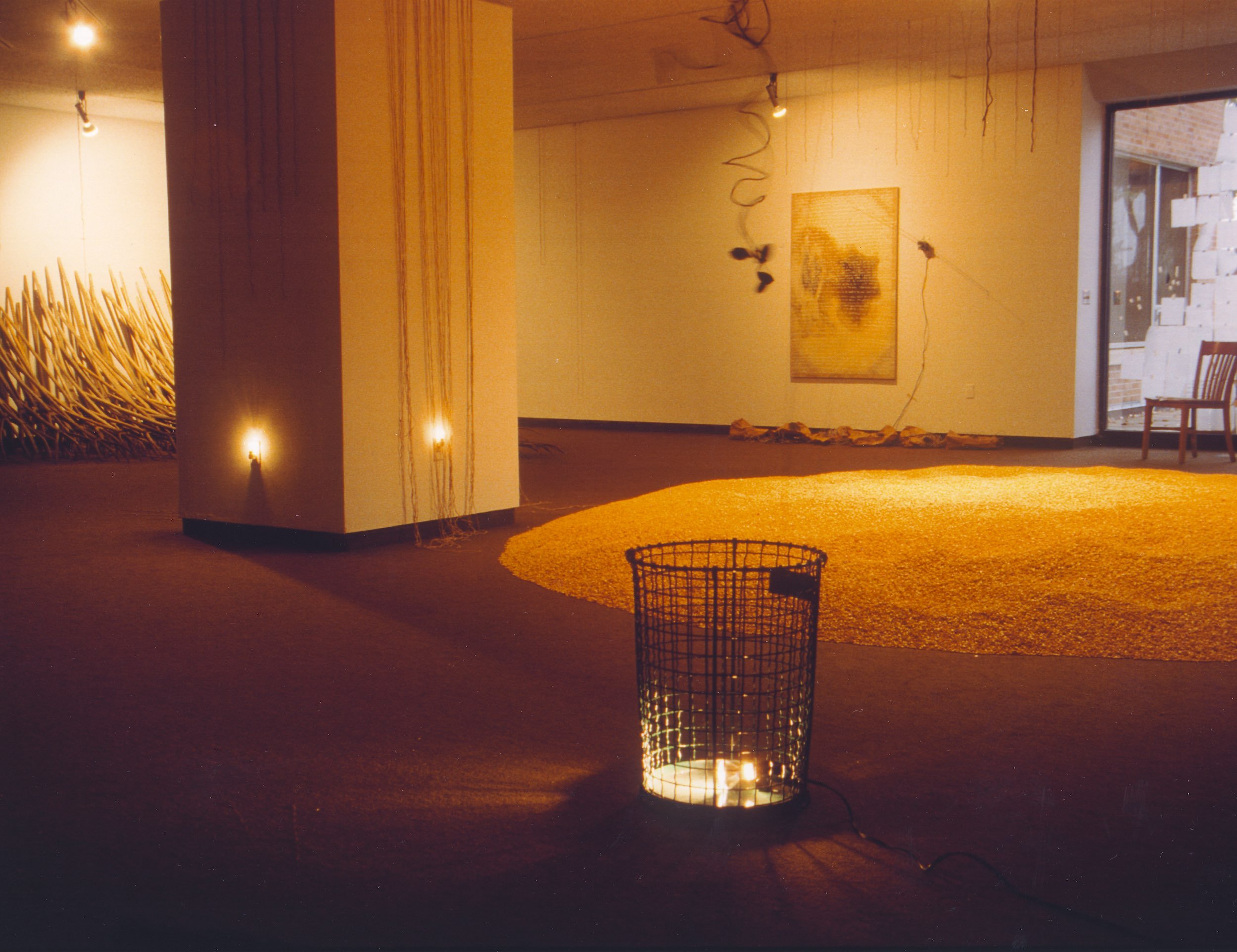 An art gallery with lighting, artwork on the walls, and decorative elements including a circular orange rug, a patterned hanging mobile, and a wire basket with a candle inside.