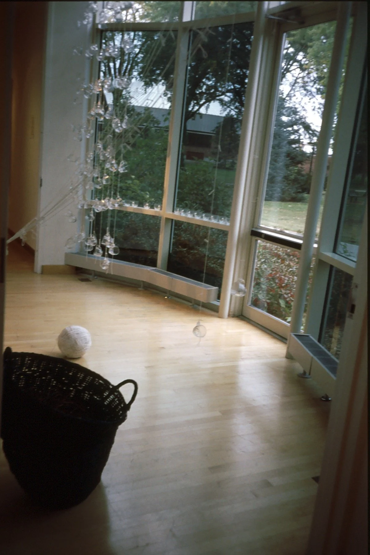 An interior view of a modern room with large floor-to-ceiling windows, hardwood flooring, a hanging decorative glass art piece, a black woven basket in the foreground, and a white ball on the floor.