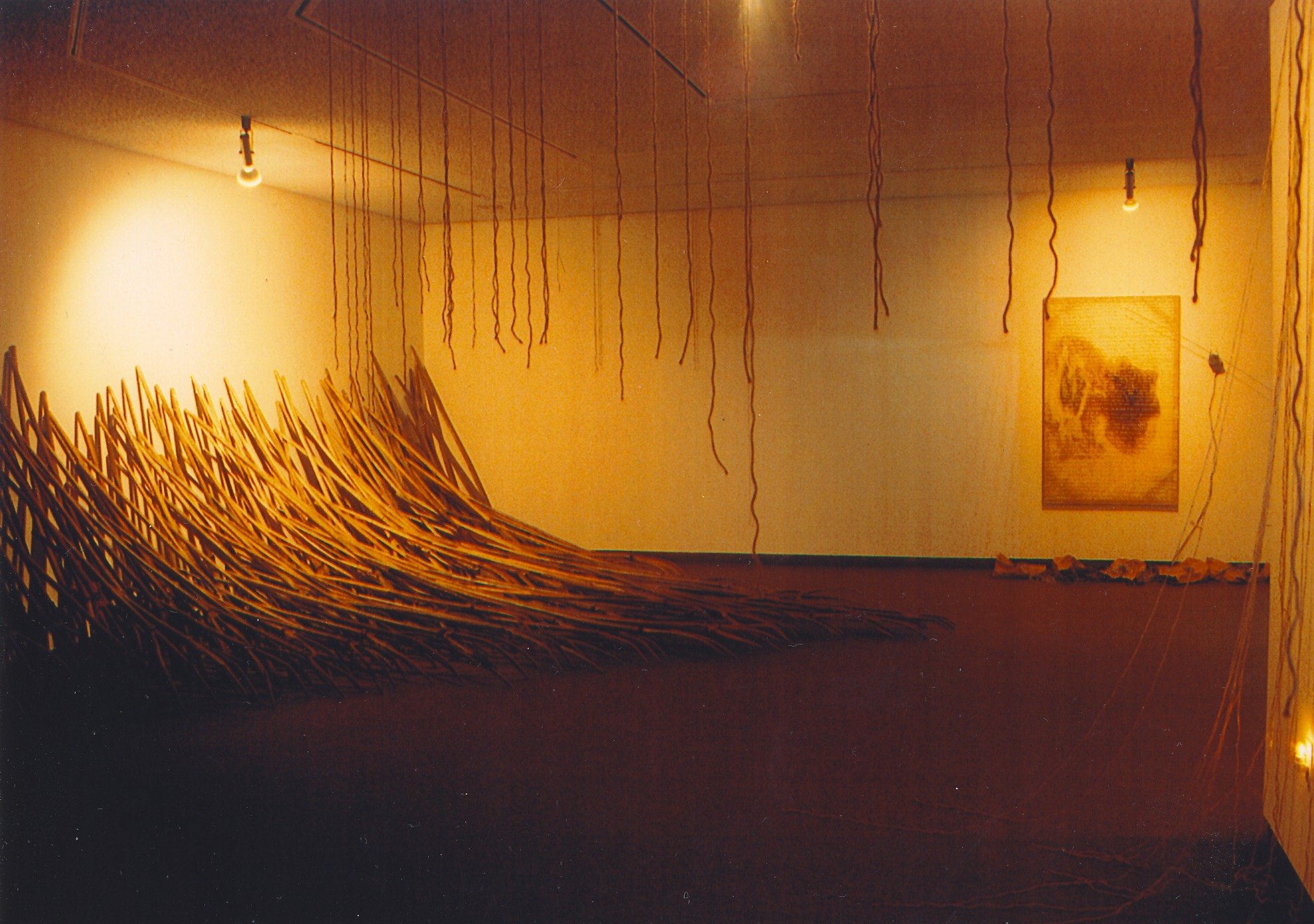 An art installation featuring a large, curved wooden sculpture on the floor, suspended hanging rods from the ceiling, and a wall painting in the background.