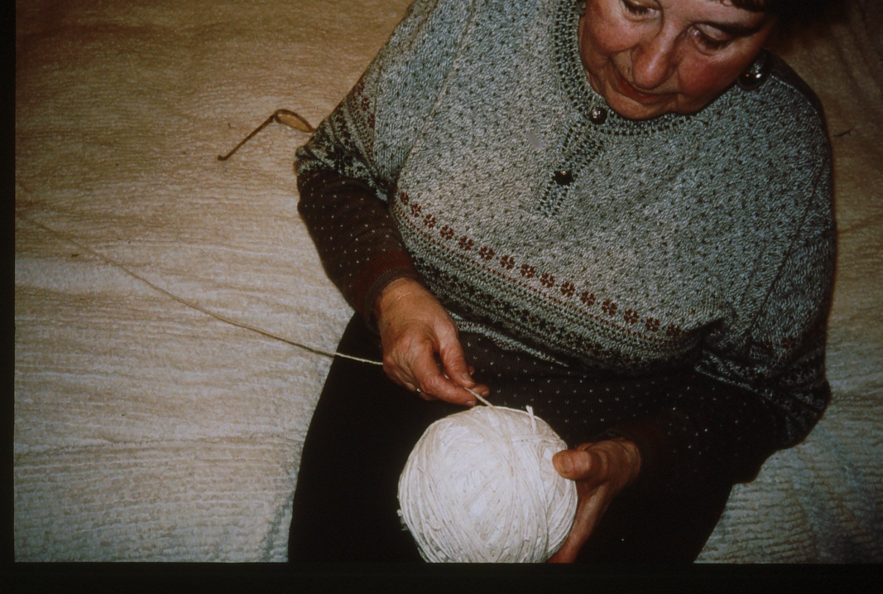 An elderly woman knitting with white yarn, sitting on the floor.
