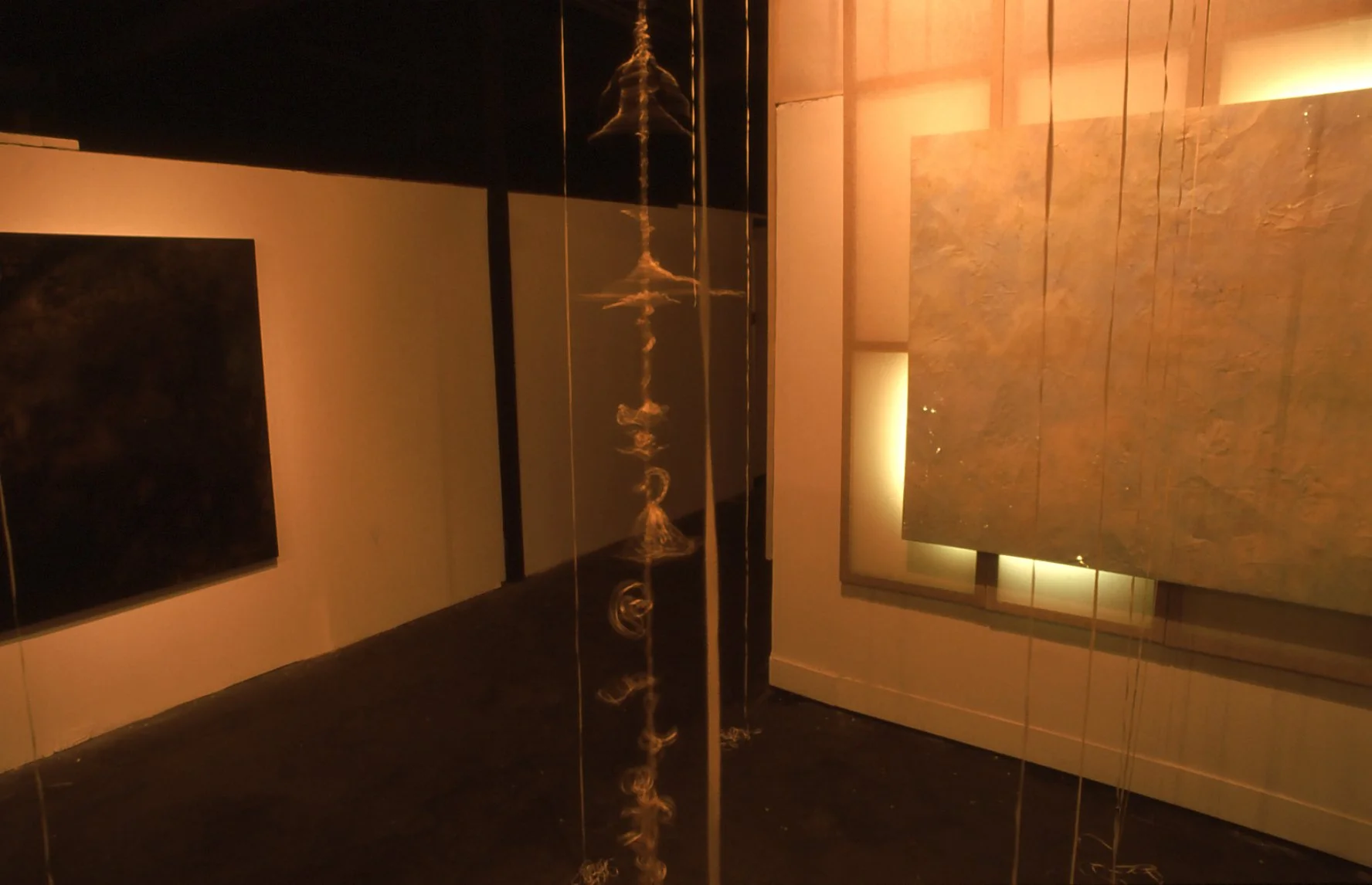 An interior view of an art installation featuring a vertical gold-colored sculpture or structure with swirling, cloud-like patterns glowing softly, surrounded by square panels with textured art pieces and warm lighting.