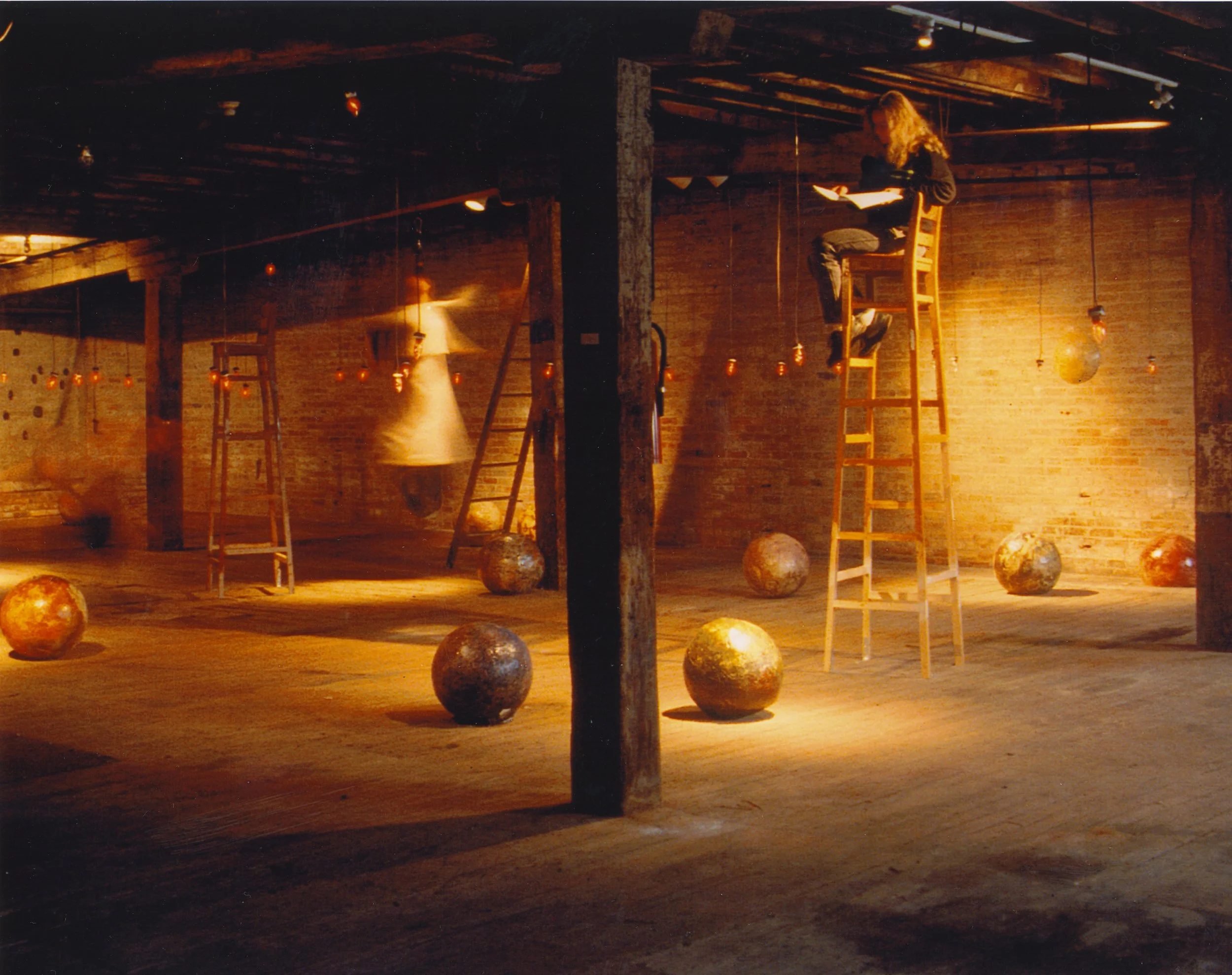 An indoor art installation with multiple ladders, floating spheres, and a person reading on a high ladder in a dimly lit room with wooden walls and brick pillars.