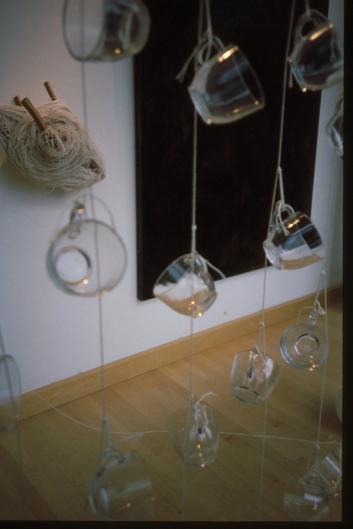 Glass cups hanging in a decorative arrangement in front of a dark panel, with a shadow of a ball of yarn on the wall to the left.