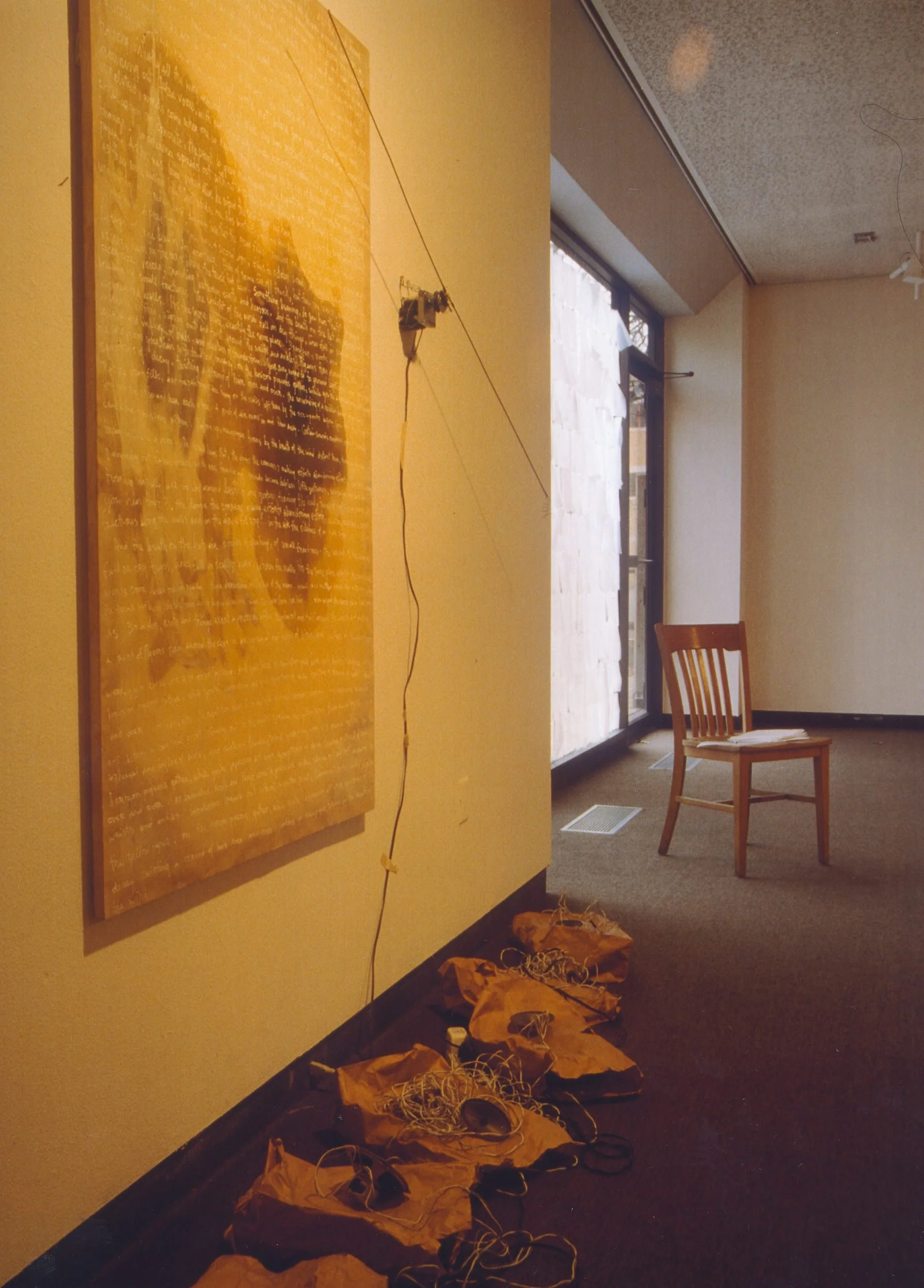 An art installation on a wall featuring a large yellow textured painting, with a string connecting to a mechanical device, and numerous crumpled brown paper bags on the floor with tangled wires. A wooden chair and window are in the background.