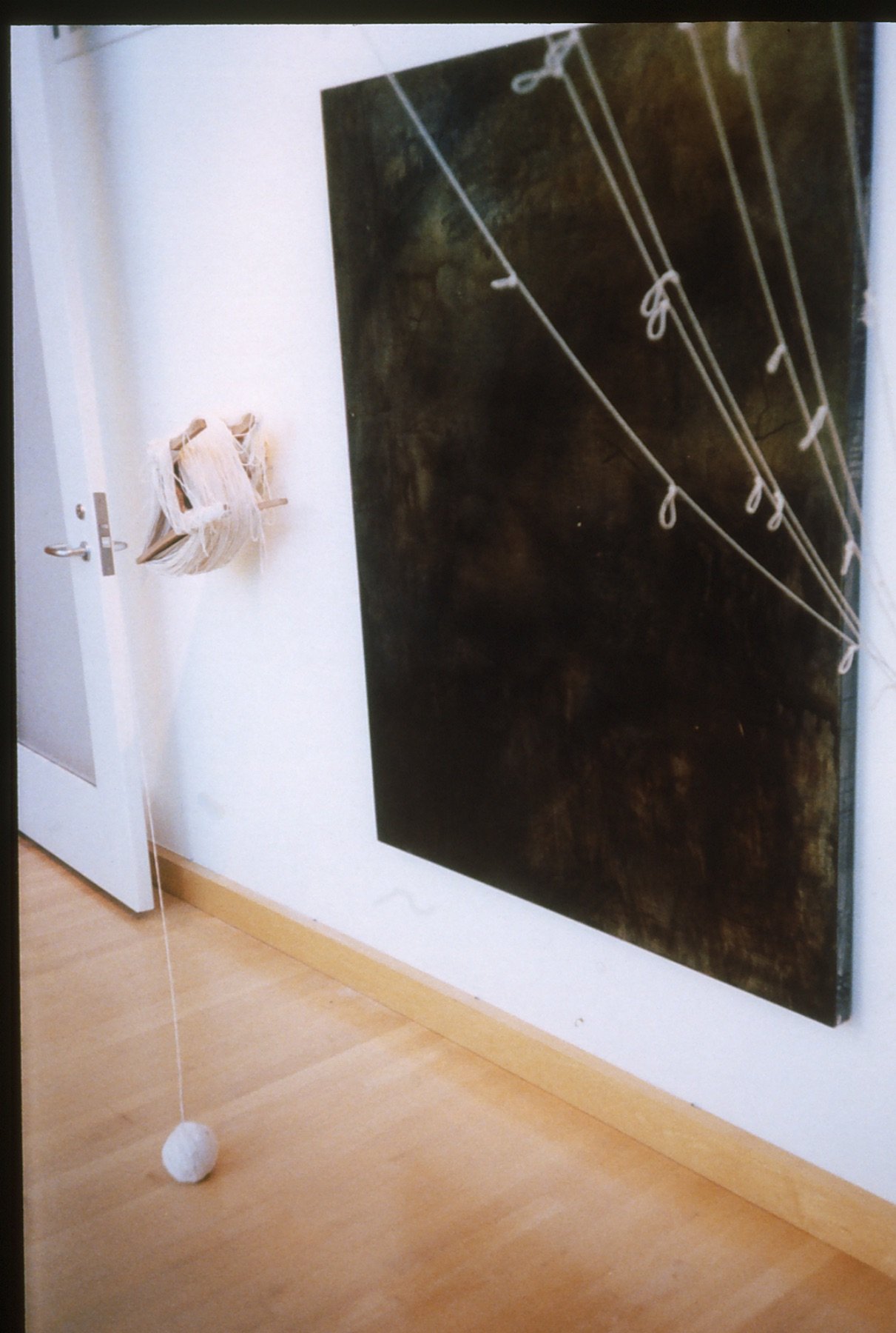An art installation featuring a large dark painted canvas with white strings tied in knots, a wooden wall hanging with strips of fabric or thread, and a string hanging from the ceiling with a white round object attached at the bottom, all set against