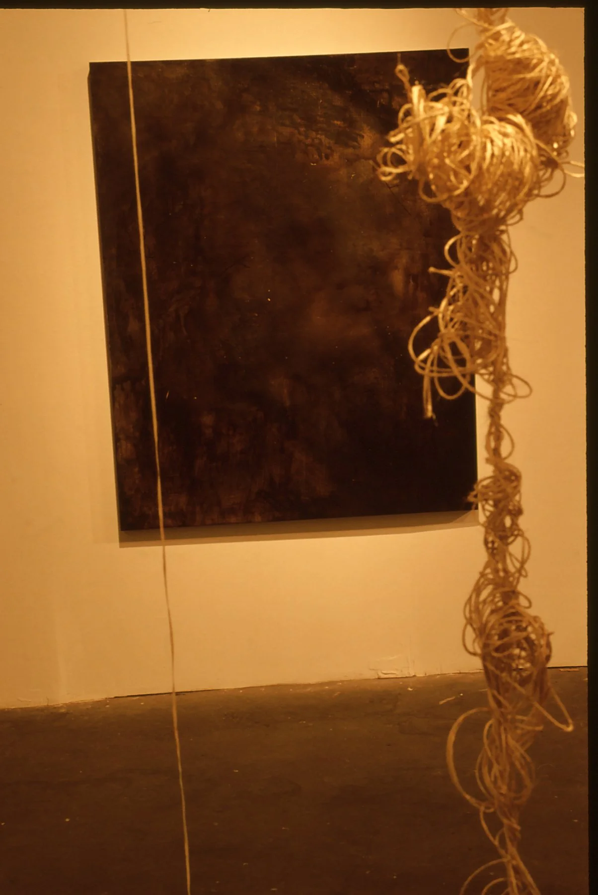 An art gallery features a large, dark, textured canvas on the white wall with a string or rope hanging vertically in front of it. There is a cluster of tangled, beige-colored material to the right of the canvas.