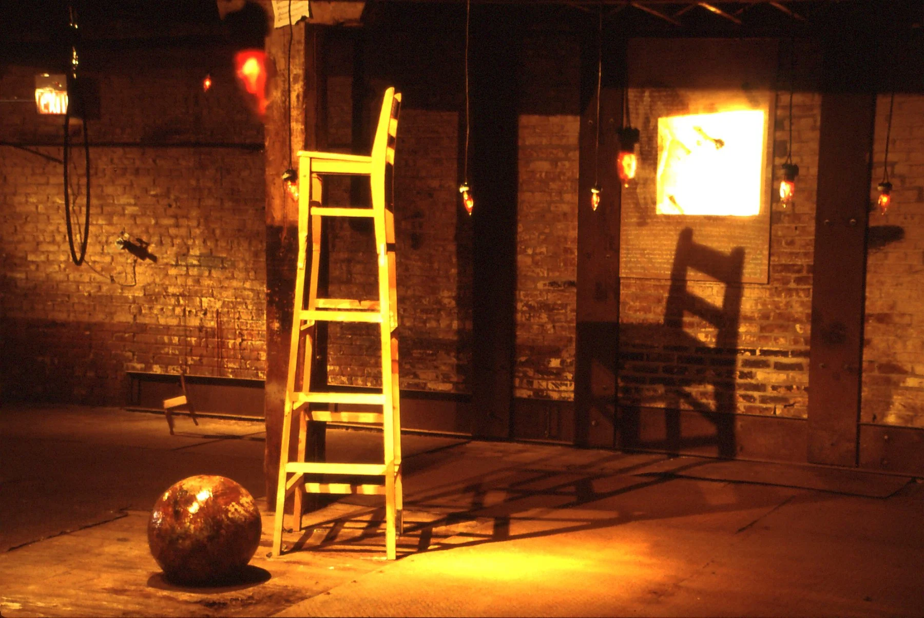 An empty room with a brick wall, illuminated by warm orange lighting, contains a yellow wooden step ladder, a reflective spherical object on the floor, and a shadow of the ladder cast on the wall.