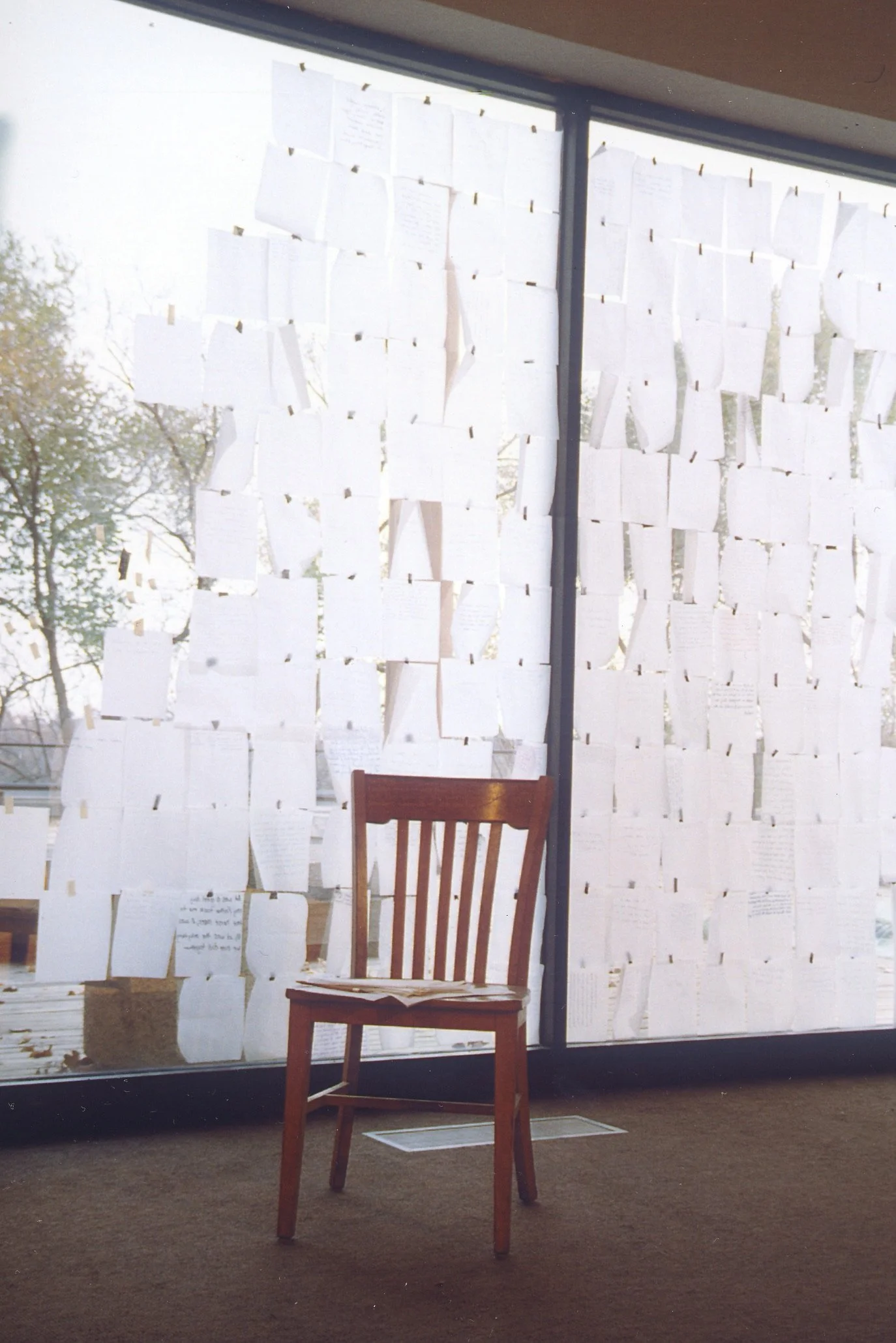 A wooden chair placed indoors in front of a large window covered with numerous pieces of paper attached with clothespins, with trees visible outside.