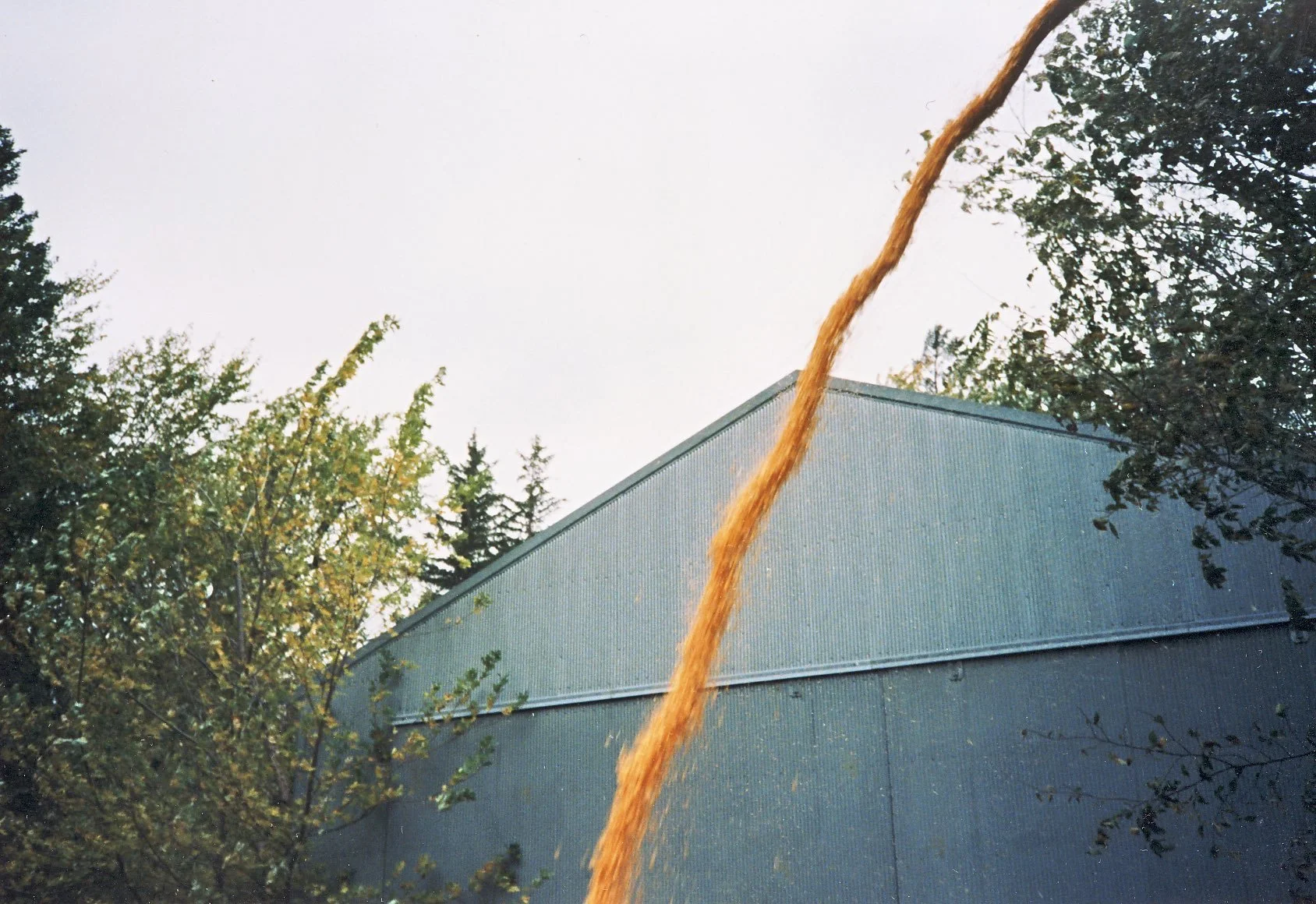 A stream of orange liquid pouring downward near a blue industrial building surrounded by trees.