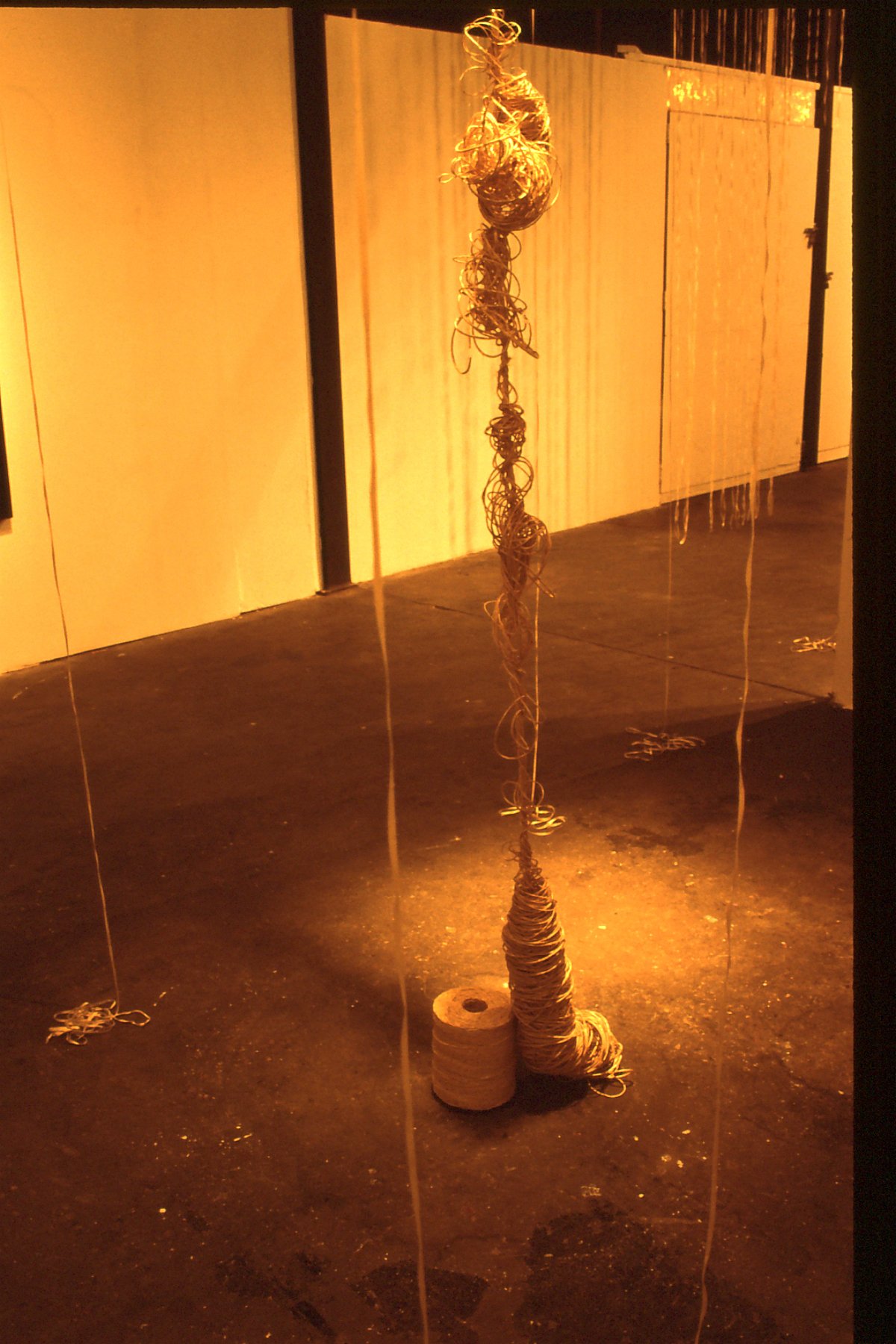 An art installation featuring a vertical arrangement of wire loops and coils, supported by two stacked rolls of tape on the floor, illuminated by warm yellow lighting.