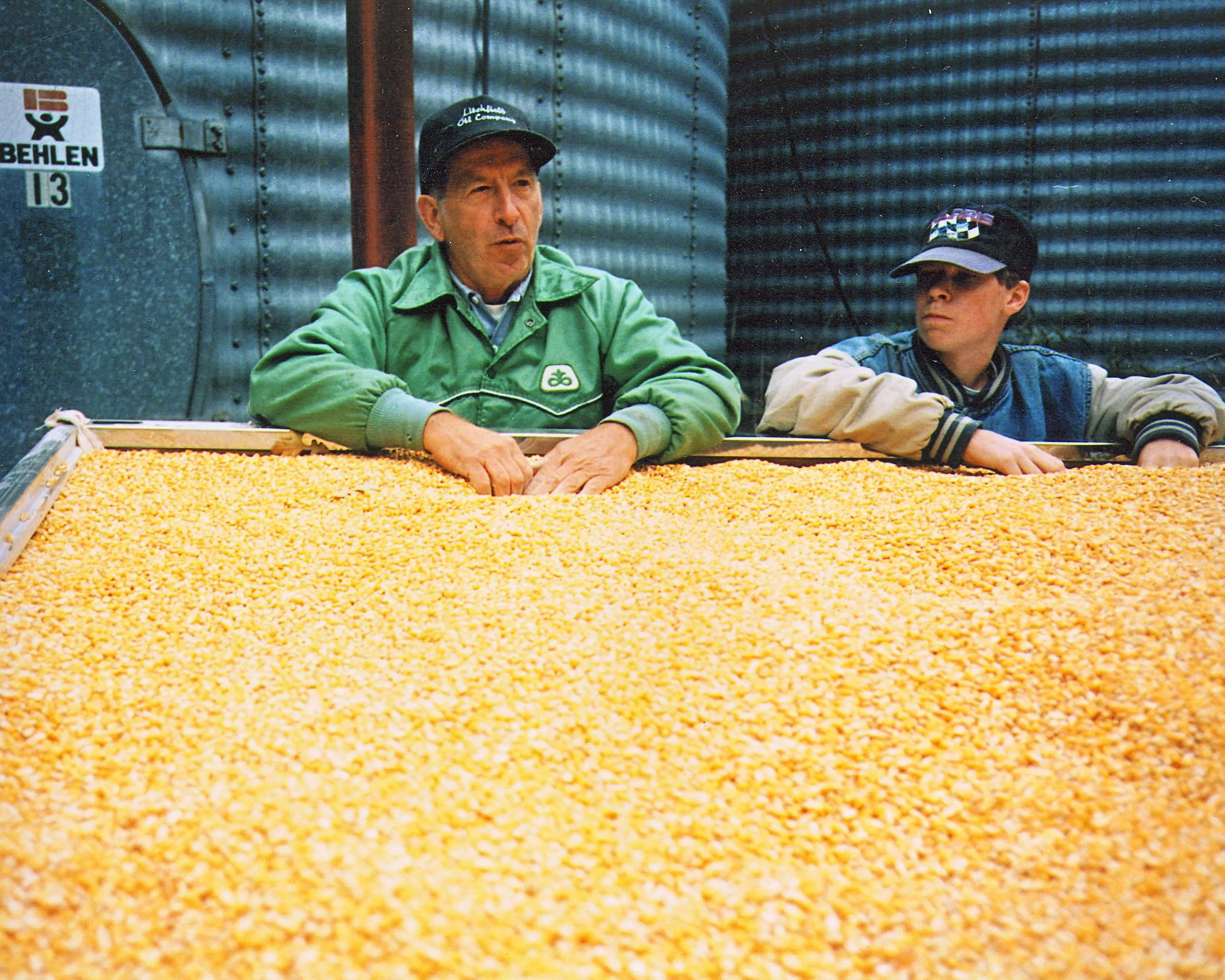 Two men, one older and one younger, standing behind a large pile of yellow corn. The older man wears a green jacket and a black cap, while the younger man wears a denim jacket and a black cap. They are in an industrial setting with metal silos in the