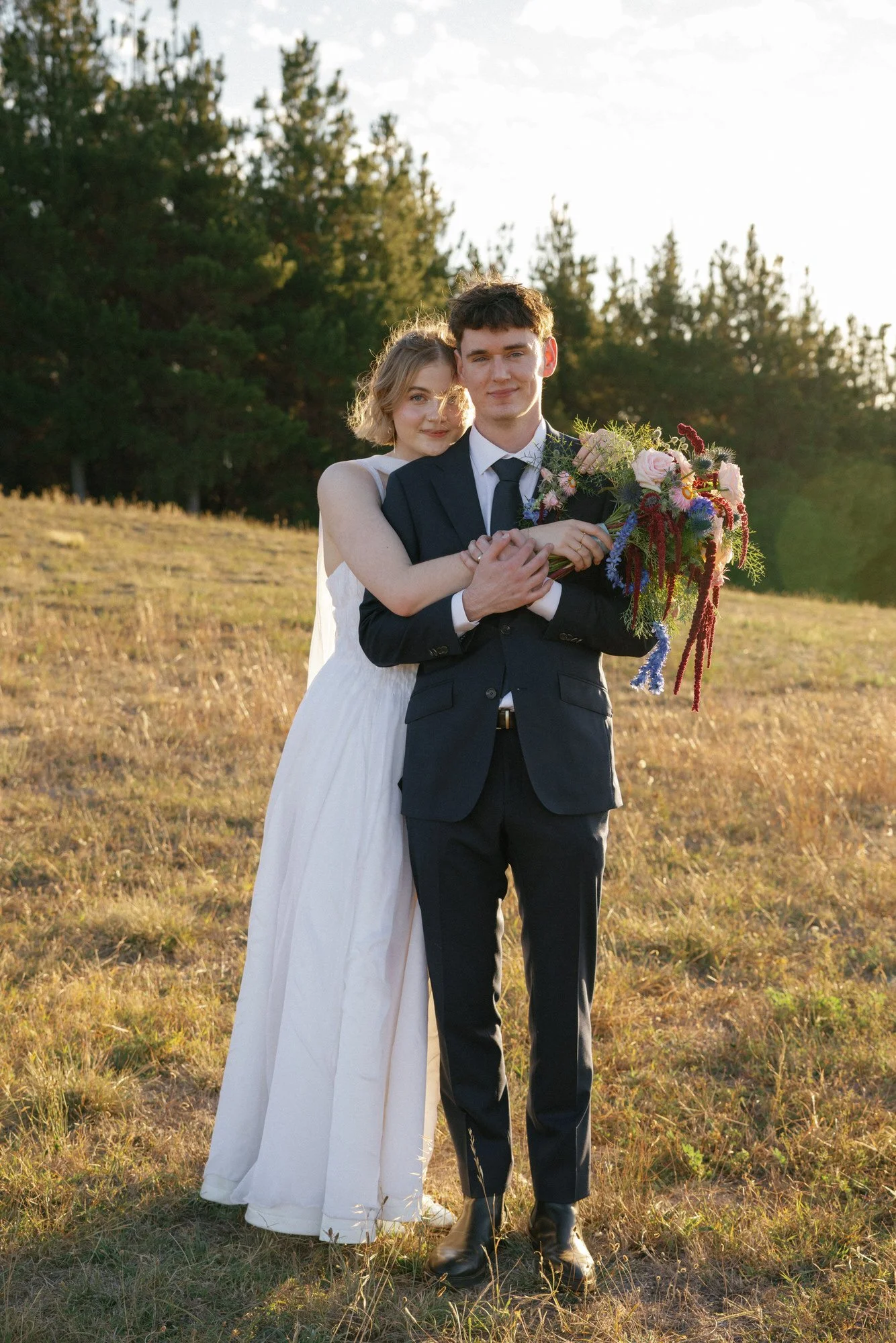 Newlyweds embrace in golden afternoon light against a backdrop of tall pines, bouquet spilling with soft pinks and wild textures