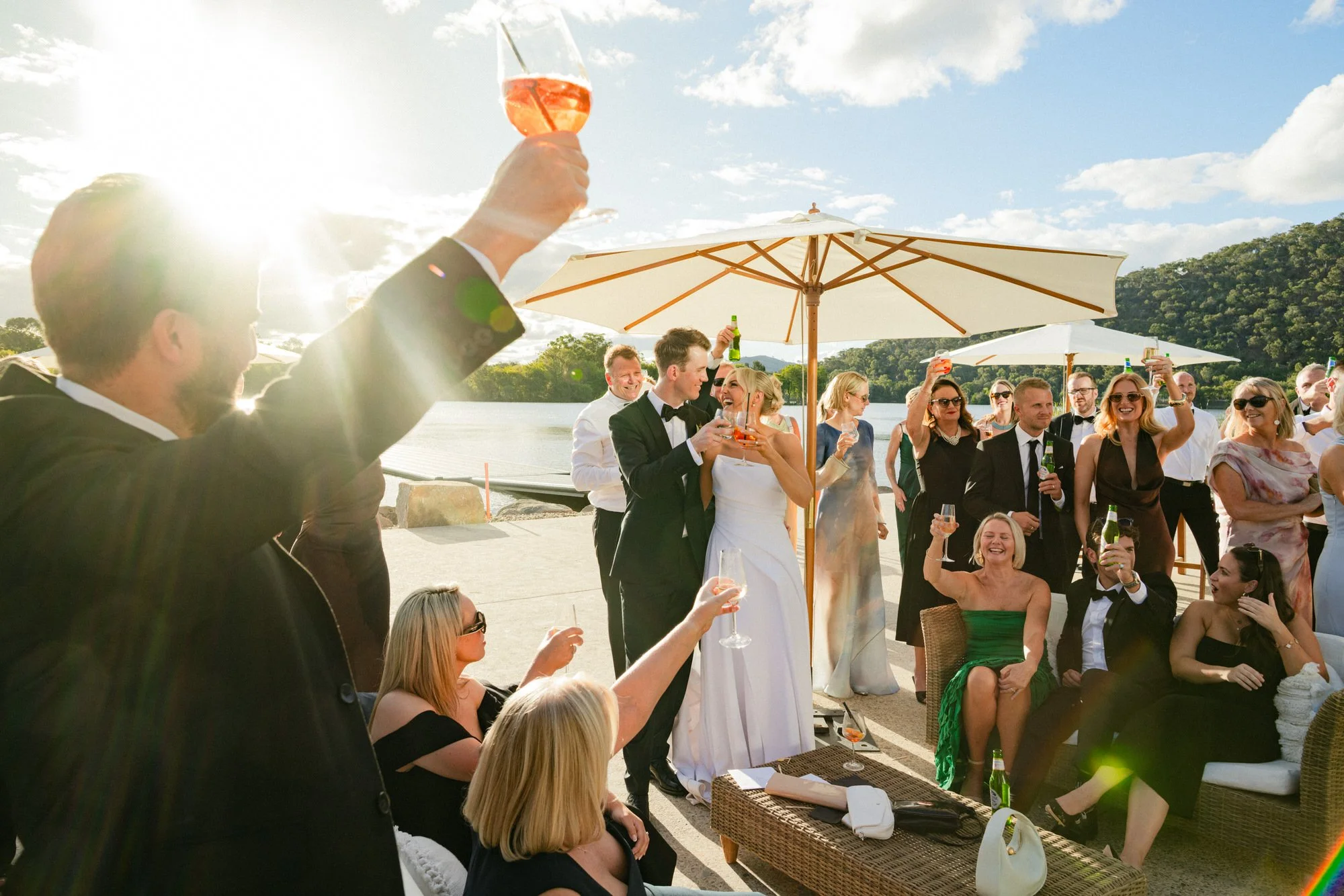 Newlyweds share a toast with guests during golden hour by the lake at the Red Shed wedding venue in Canberra, with champagne glasses raised under umbrellas.