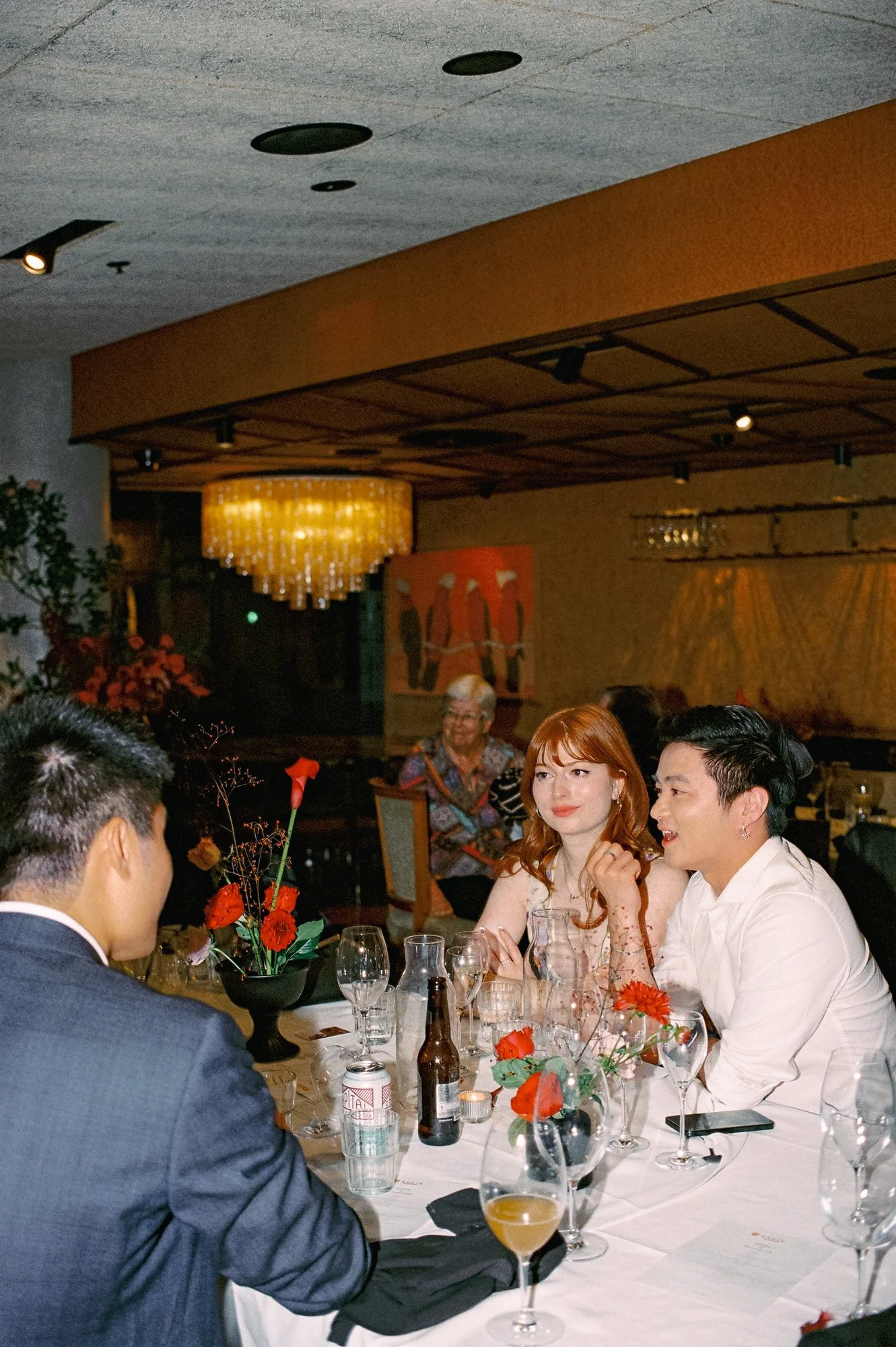 Guests talking and drinking at a wedding reception at Monster Kitchen & Bar, surrounded by red floral arrangements and mid-century interiors. 35mm film photograph with a warm glow