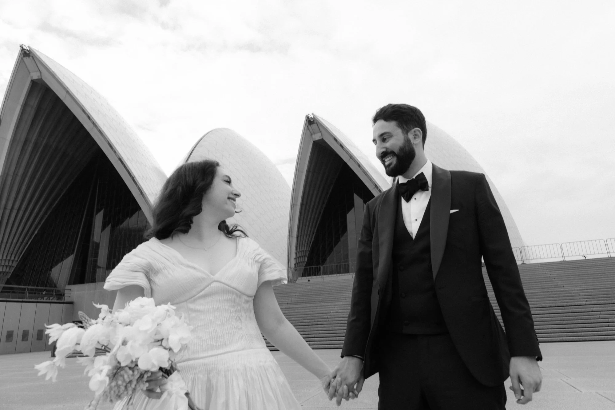Bride and groom holding hands and smiling at each other in front of the Sydney Opera House sails, captured in timeless black and white.