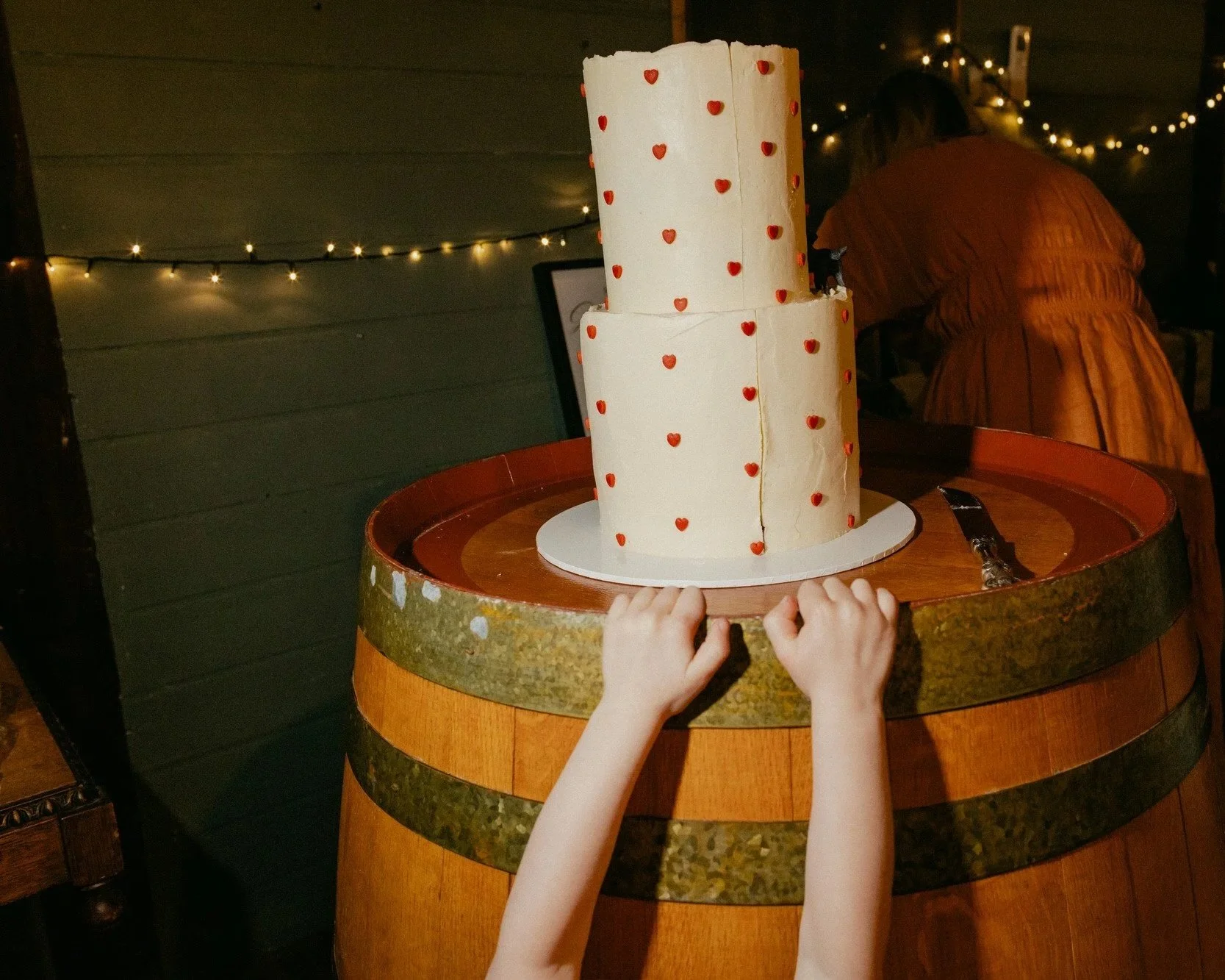Child reaching up for a whimsical two-tier wedding cake decorated with red hearts on a wine barrel at a rustic Canberra wedding reception, captured by Corinna & Dylan, candid wedding photographers.