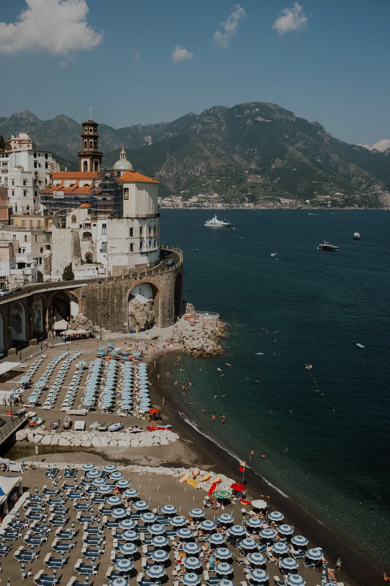 Rows of blue-and-white umbrellas line a busy beach on the Amalfi Coast, with pastel buildings perched above and mountains across the sea.