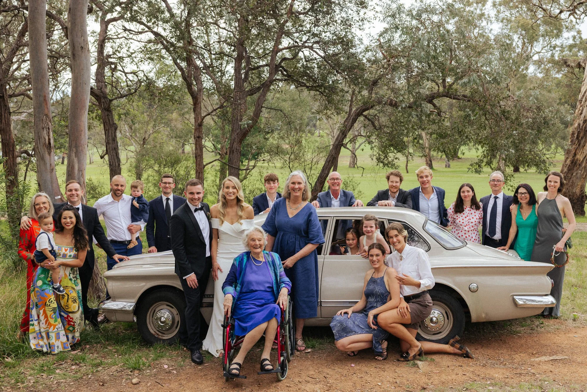 Multi-generational family portrait with bride, groom, and loved ones gathered around a vintage car at a relaxed backyard wedding near Canberra, photographed by Corinna & Dylan, candid wedding photographers