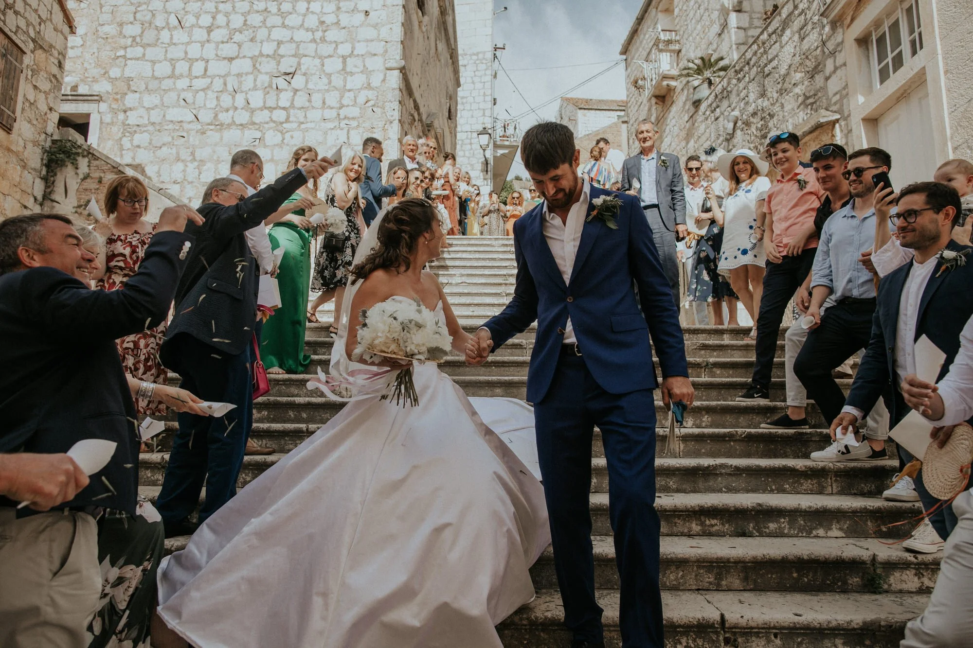 Bride and groom walk hand in hand down stone steps in Vis, Croatia, as guests cheer and toss confetti, the celebration spilling joy into the narrow streets.