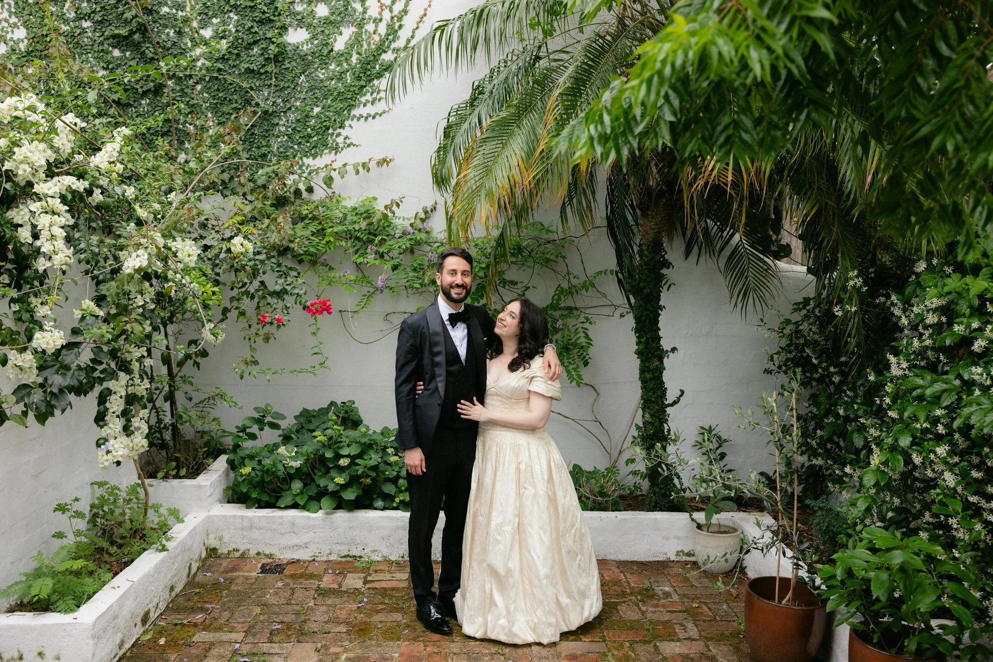 The bride and groom stand together in a leafy courtyard, framed by white walls and vines. She gazes up at him while he smiles, the scene lush and intimate.