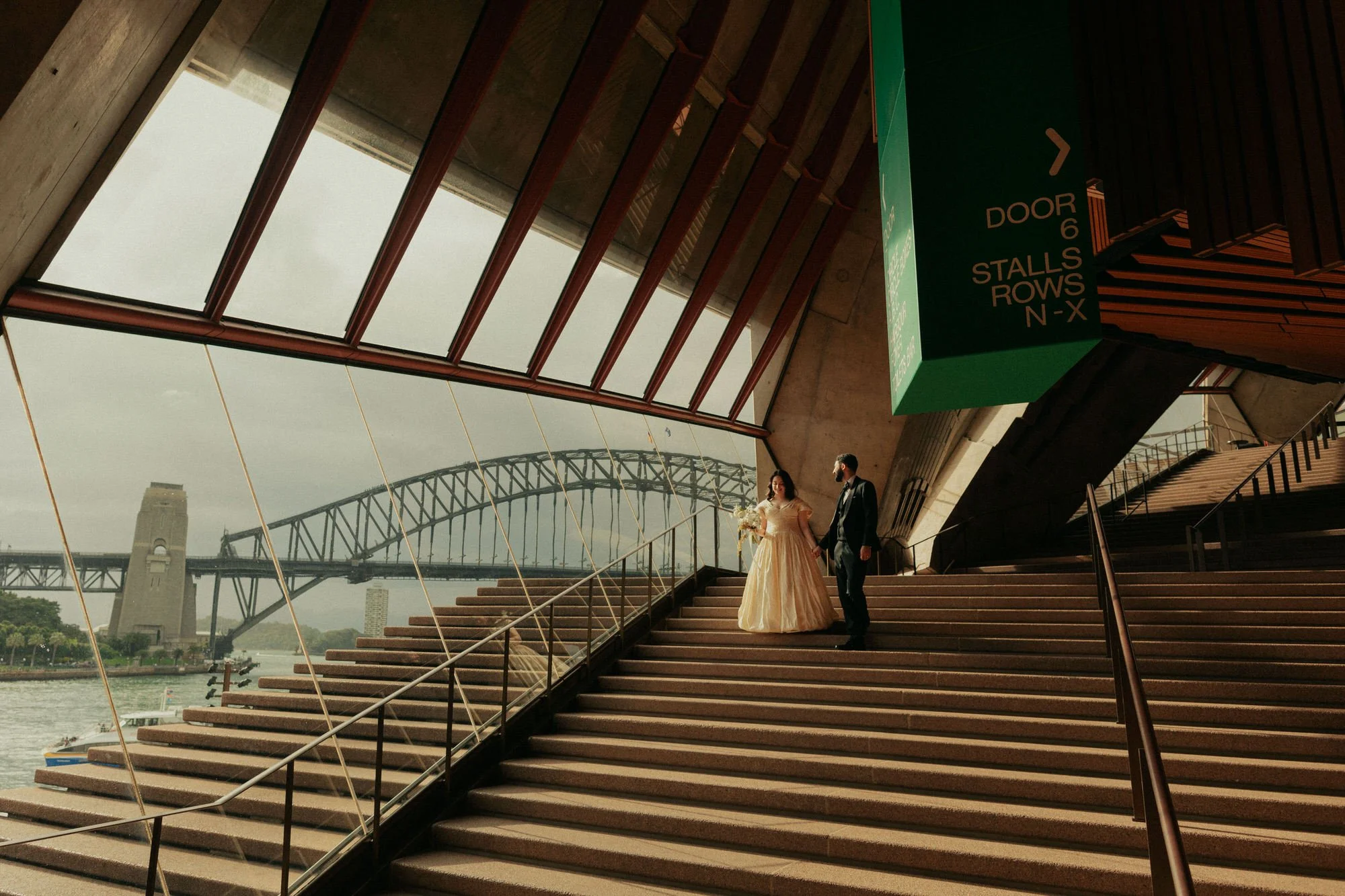 Couple framed within the soaring interior architecture of the Sydney Opera House, standing on the grand stairs with the Harbour Bridge visible beyond