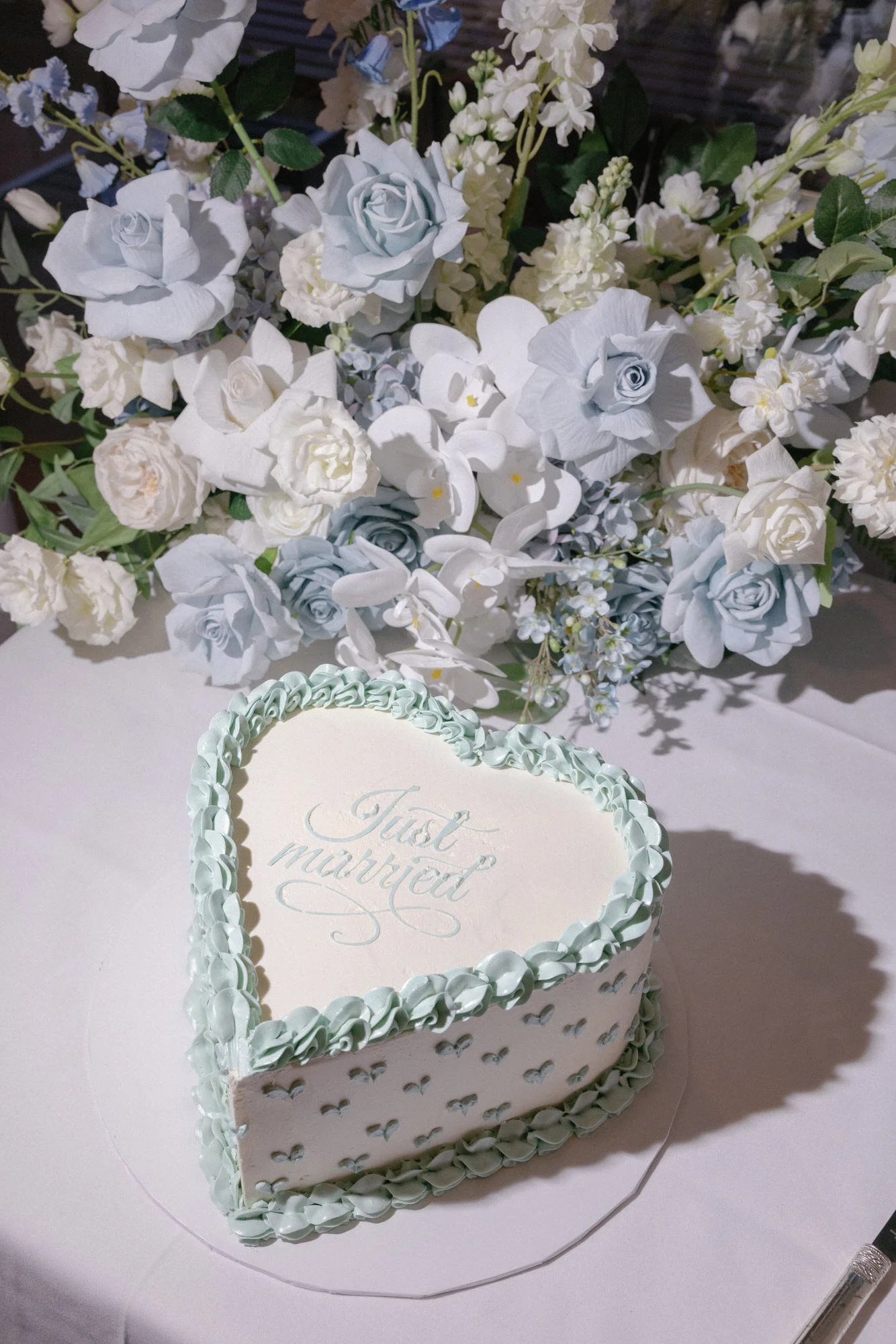 Heart-shaped wedding cake with 'Just Married' written on top, styled with pale blue icing and surrounded by pastel floral arrangements