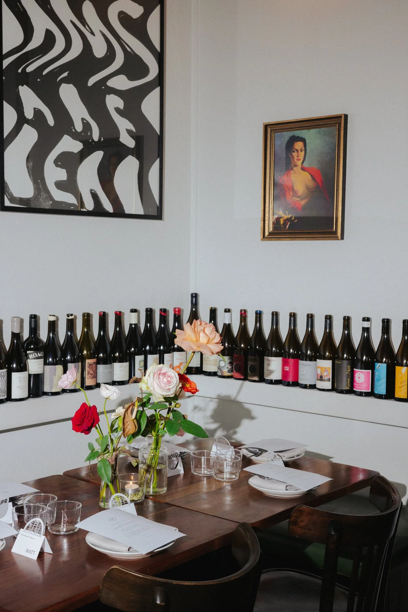 A warmly lit table set for a wedding dinner at Bar Rochford, with flowers in glass vases, neatly folded menus, and rows of wine bottles lining the wall beneath bold artwork and a vintage portrait.