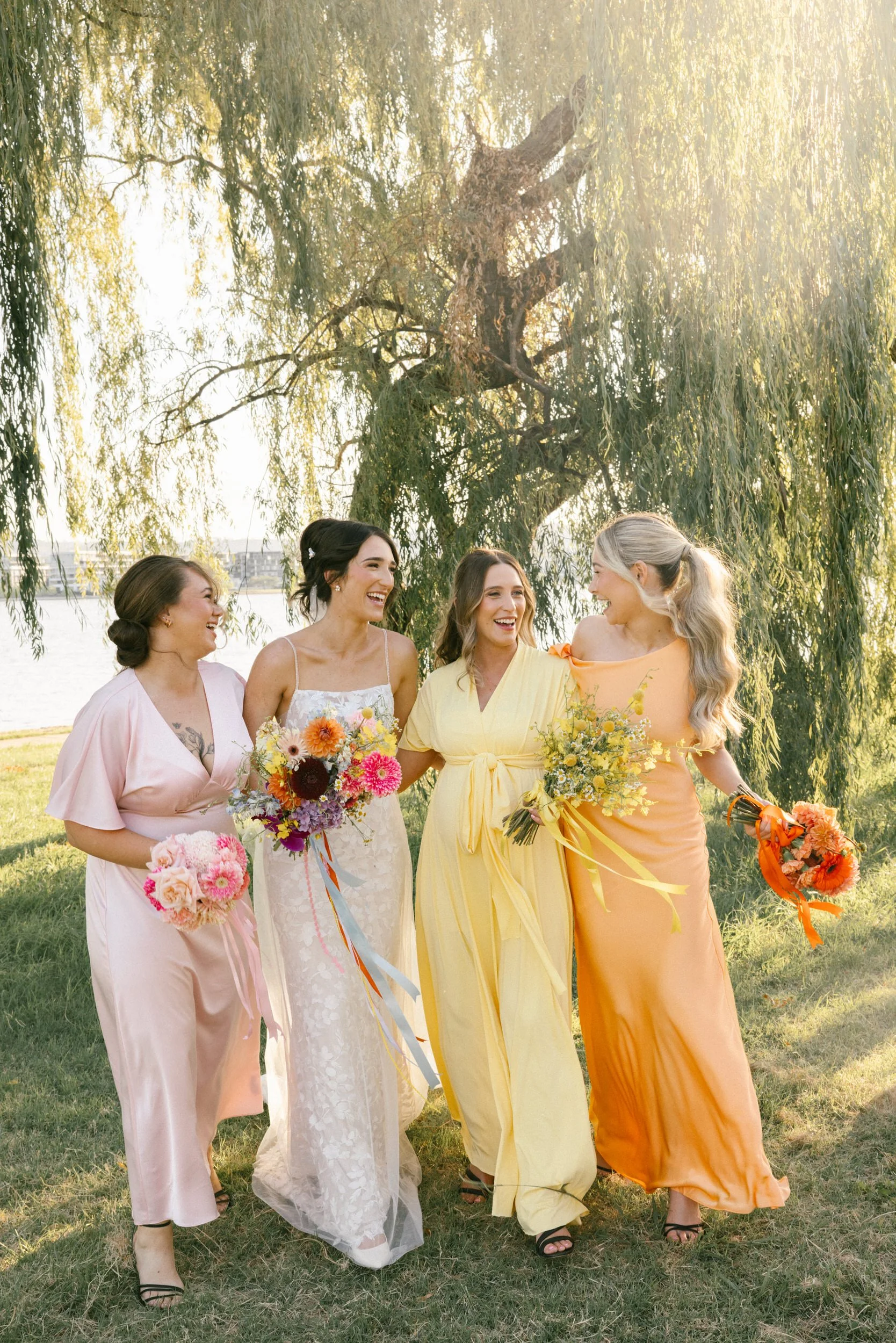 Bride laughing with bridesmaids in pastel pink, yellow, and orange gowns beneath willow trees at The Boat House Canberra wedding, captured by Corinna & Dylan, modern wedding photographers.