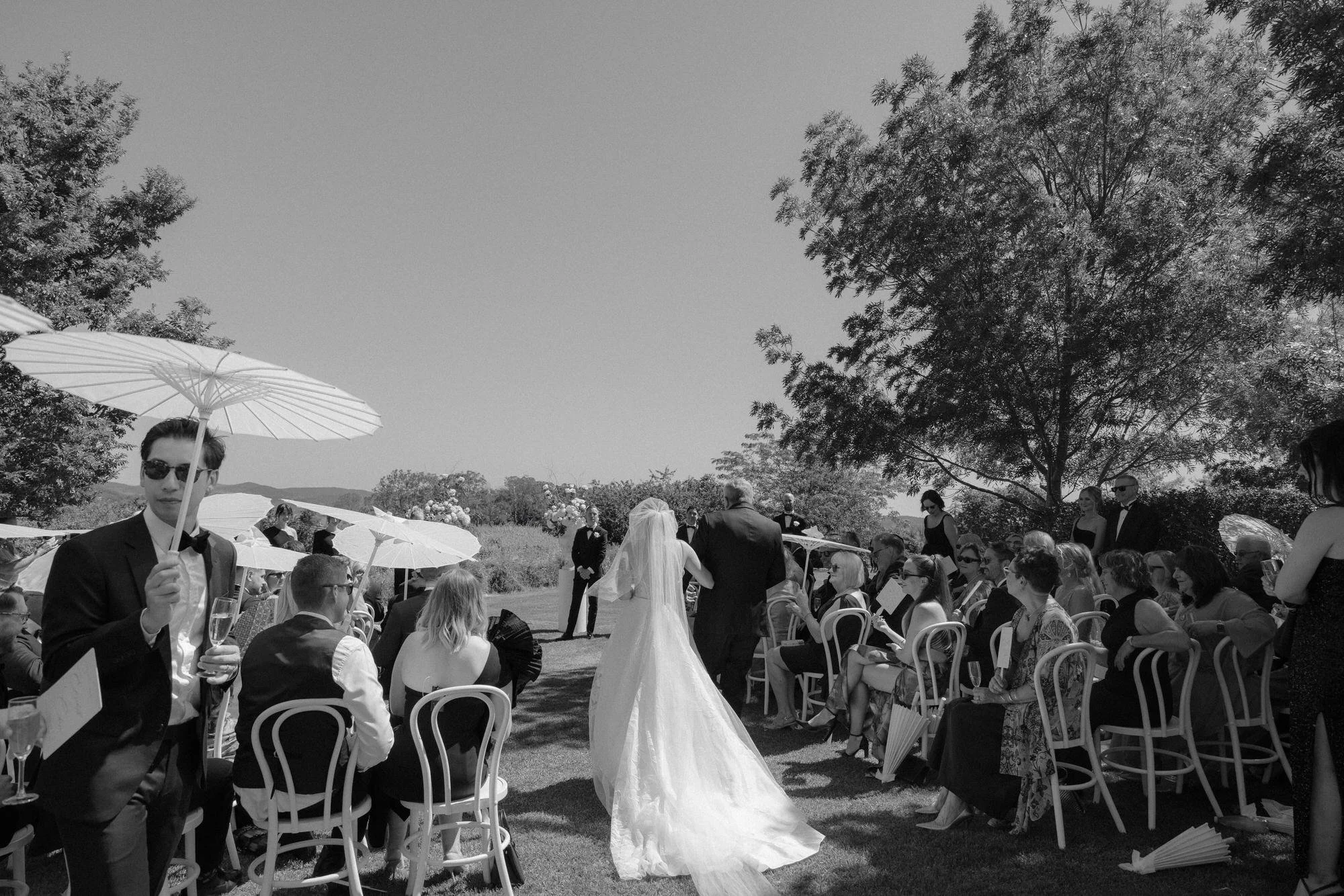 Bride walking down the aisle with her father at an outdoor Rhyanna Park wedding ceremony, surrounded by guests holding parasols, captured in timeless black and white by Corinna & Dylan