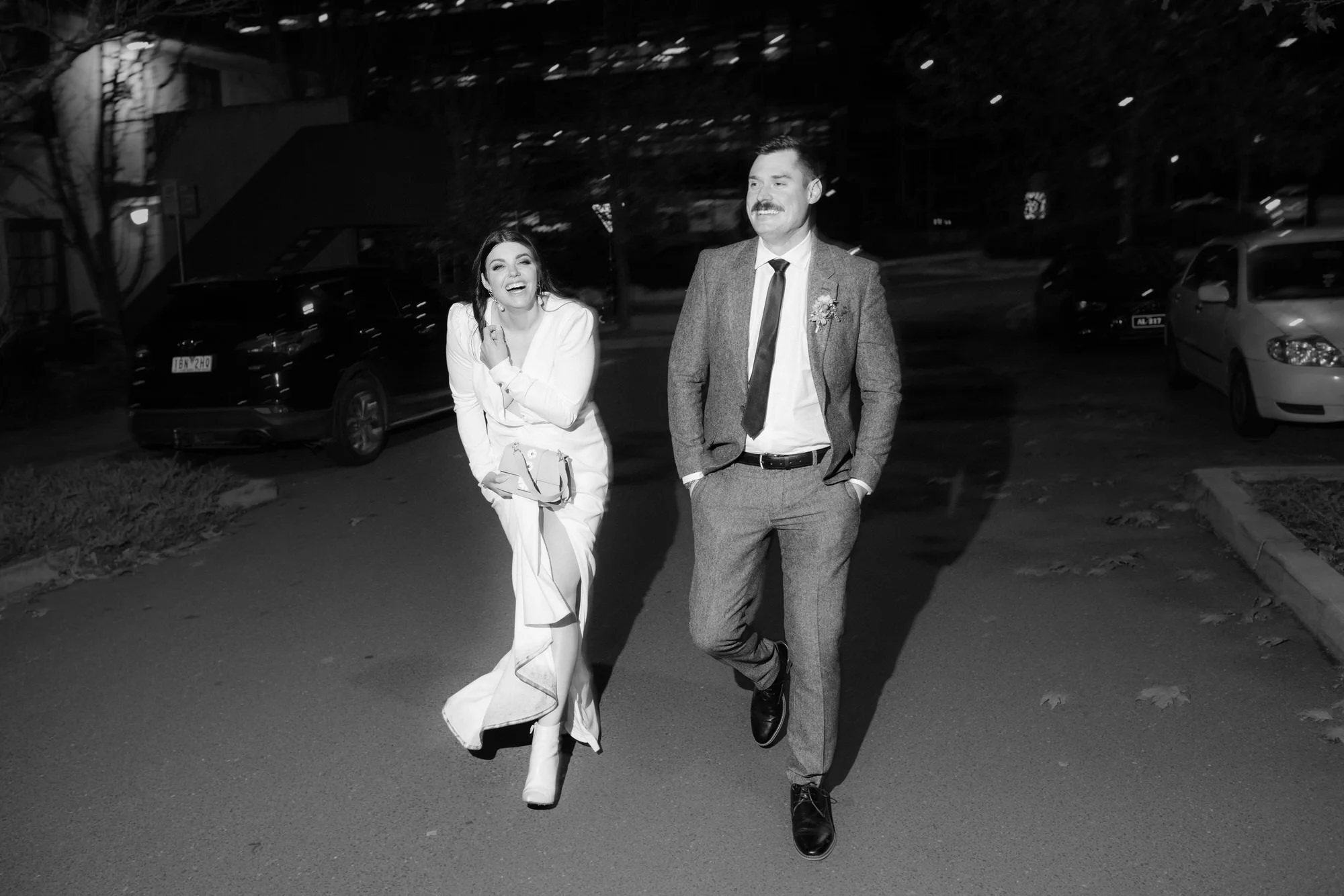 Modern Canberra elopement photography — bride in chic white suit and boots laughing with her groom as they walk through the city streets at night.