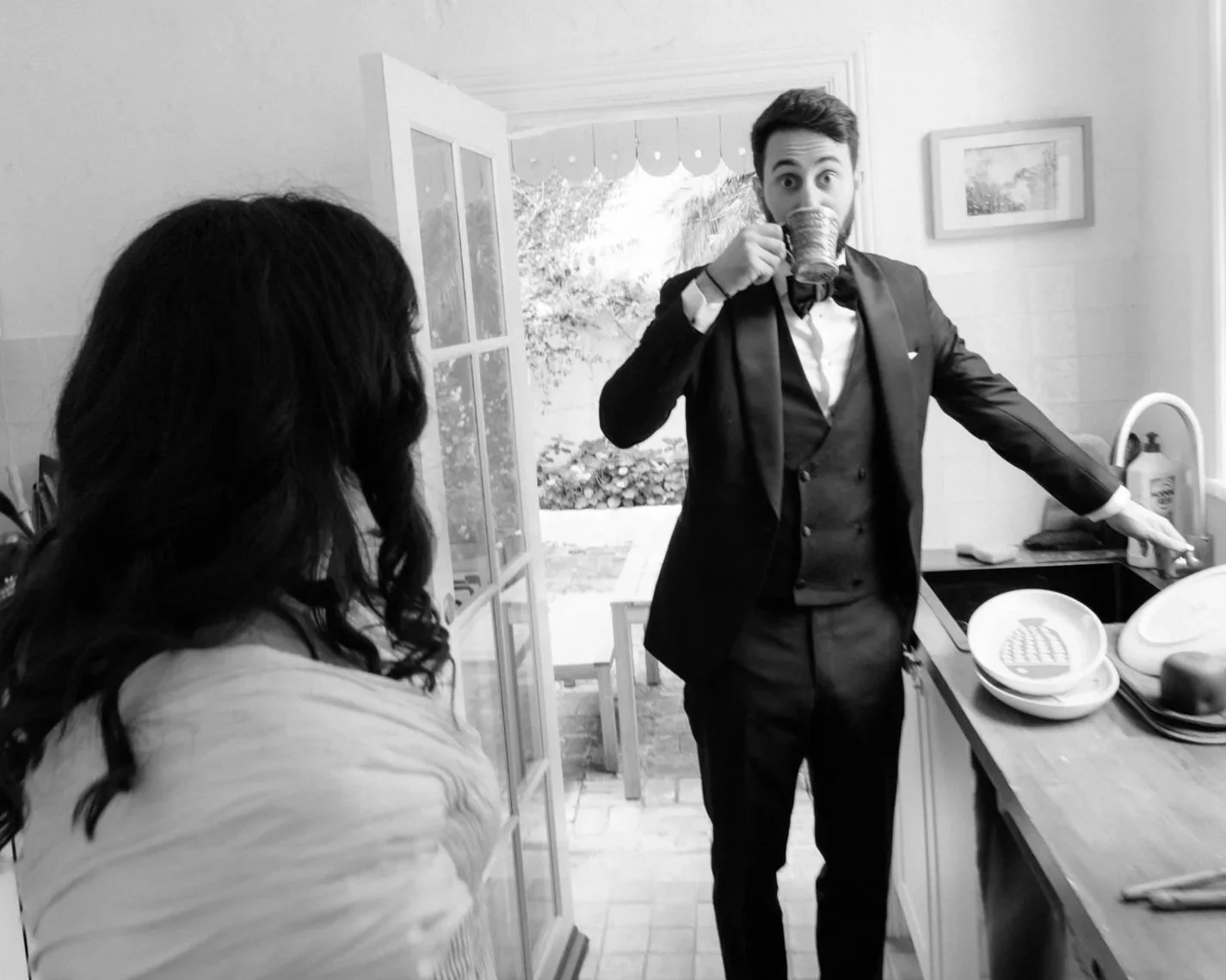 Groom in a tuxedo sips coffee in a light-filled kitchen before the wedding, caught mid-gesture in a playful, candid momen