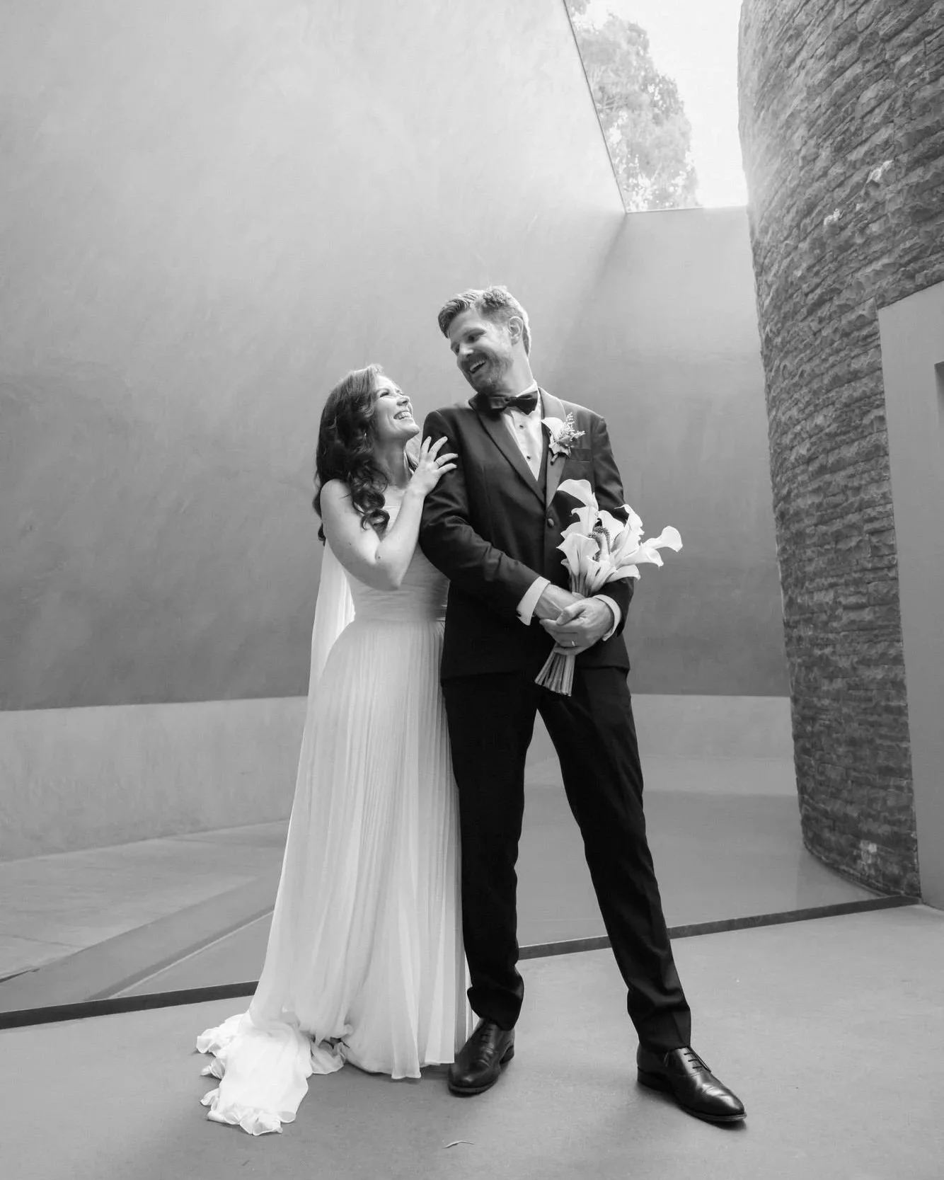 Bride and groom laugh together at the National Gallery, framed by sculptural walls, a bouquet of calla lilies in hand. Black and white, cinematic and timeless.