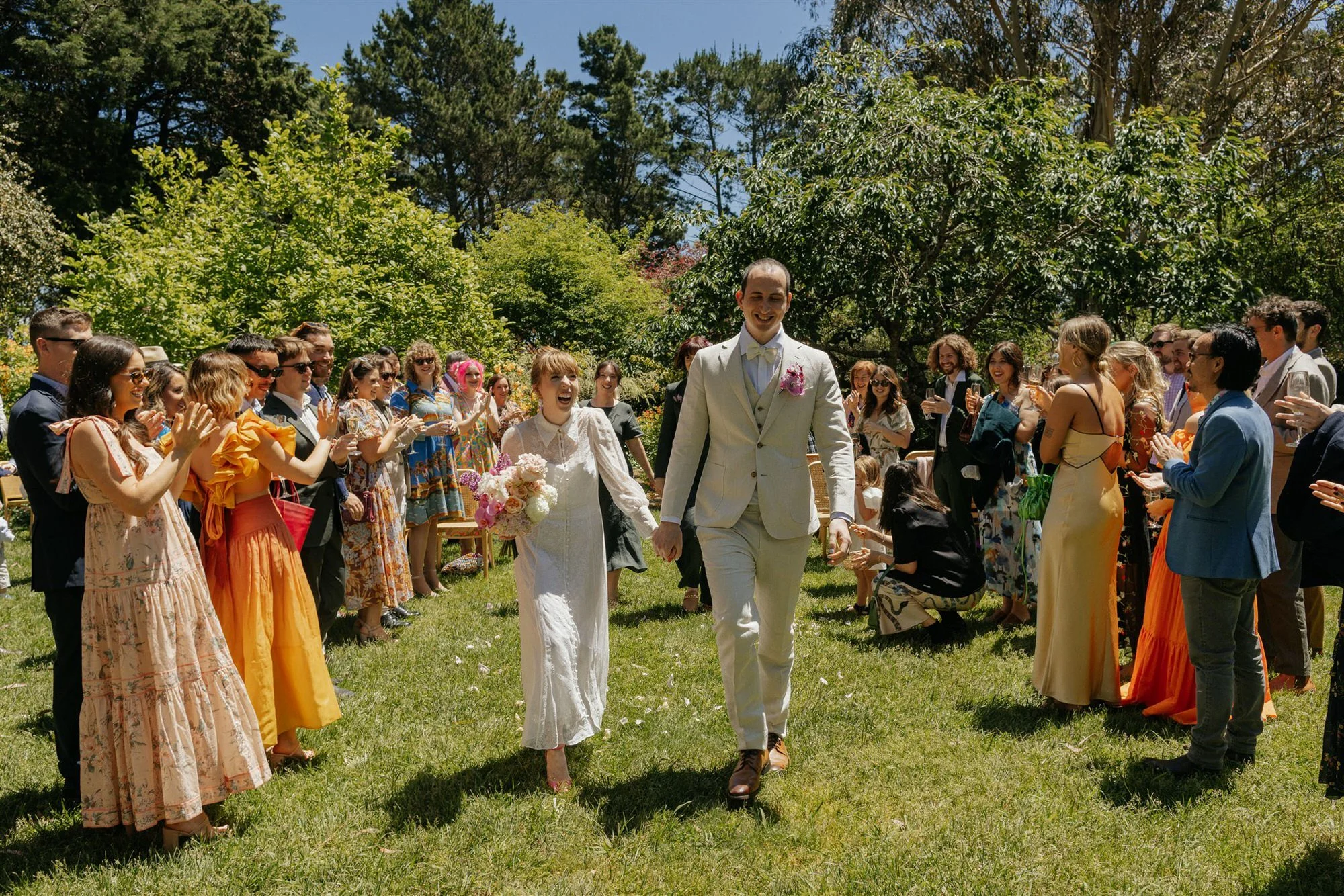 Just married couple walking back down the aisle hand in hand, laughing joyfully, surrounded by cheering guests in colorful outfits at a garden wedding in Bowral